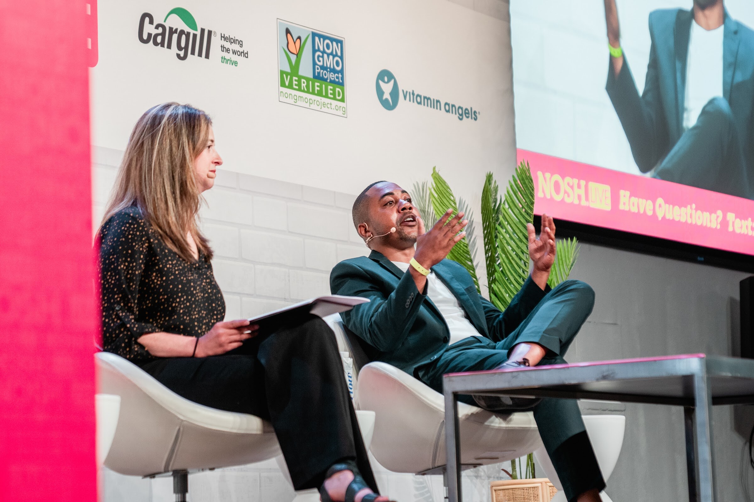 Woman with a tablet seated beside a man in a dark green blazer who is gesturing, a Nosh Live screen behind them.