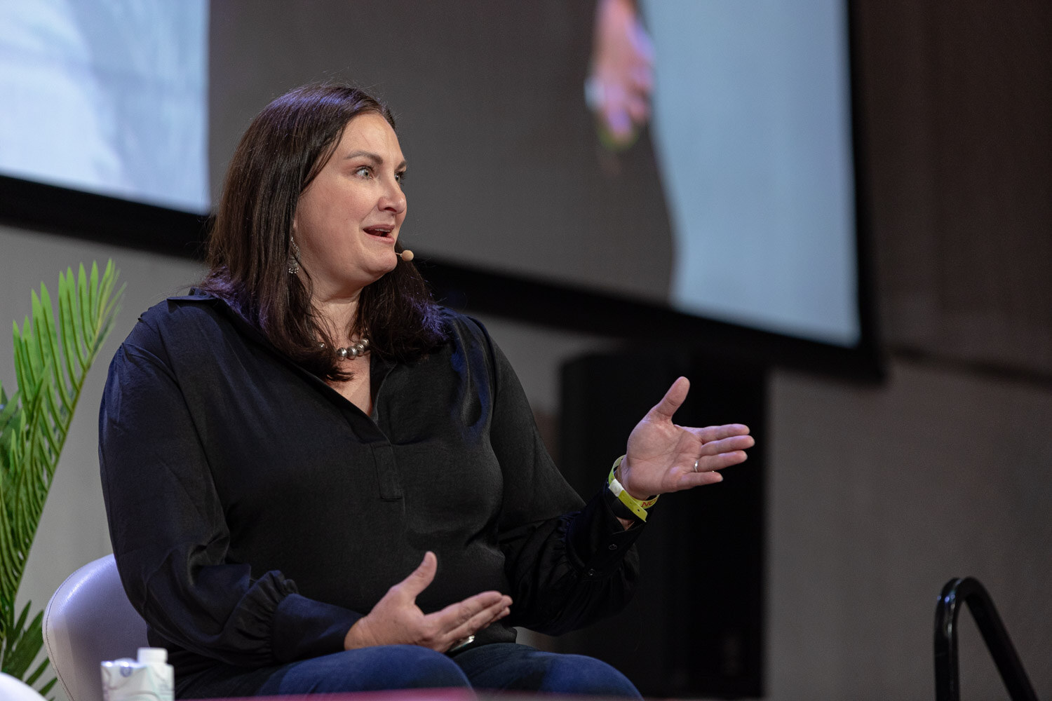 Dark-haired woman in a black blouse gesturing with one hand, a screen behind her.