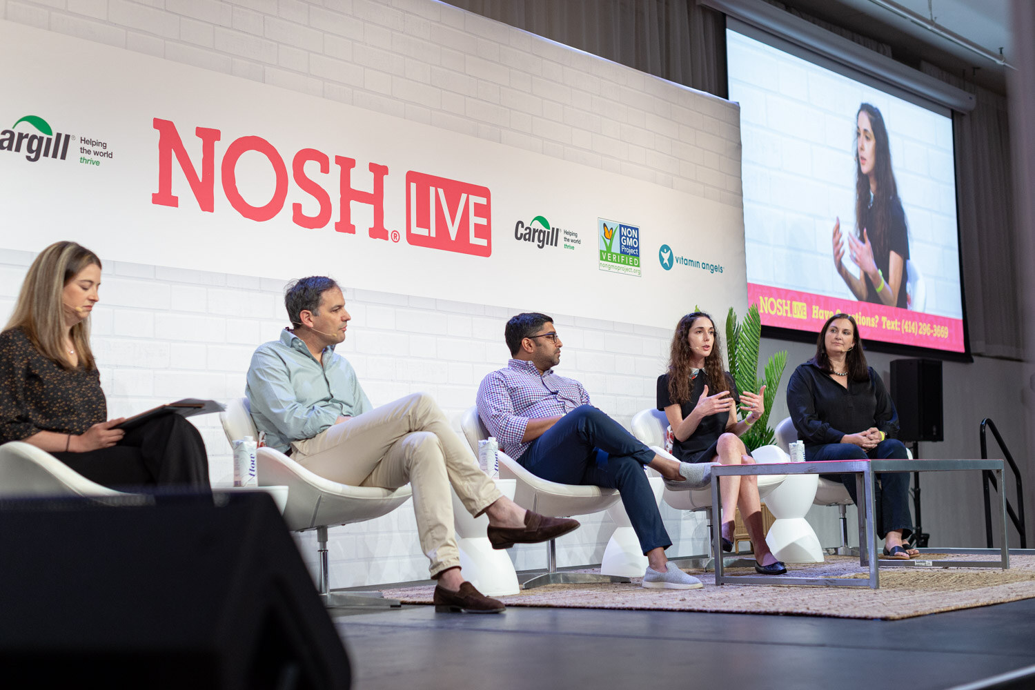 Five people seated in white chairs on stage in front of a Nosh Live wall, with a screen at the right.