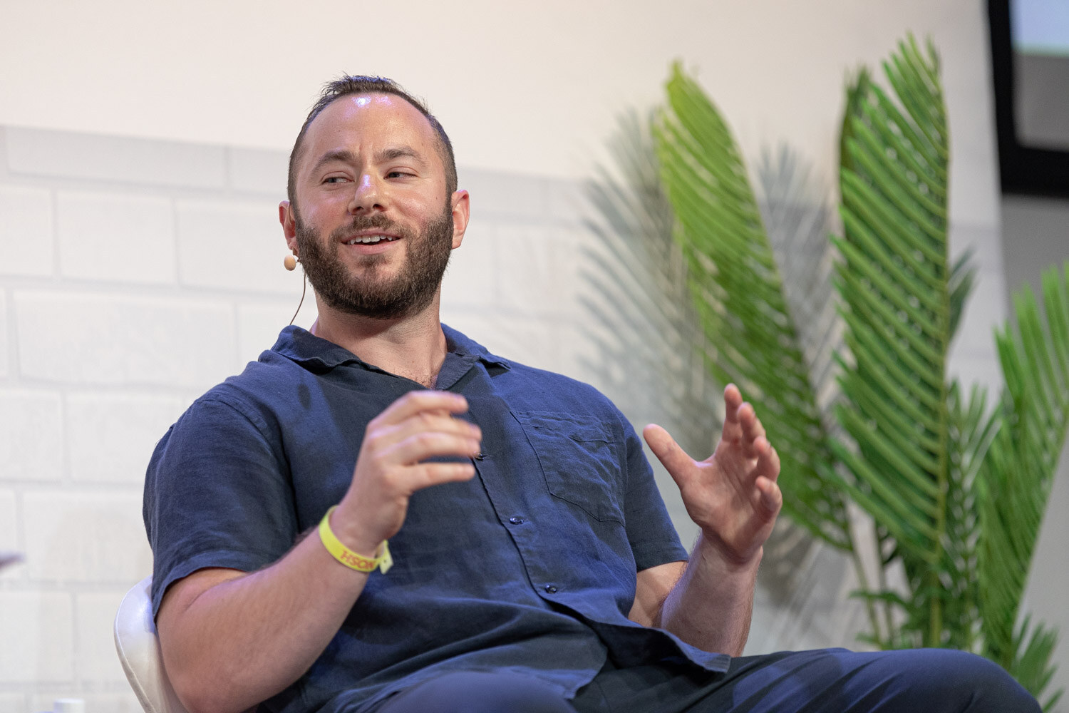 Bearded man in a navy button-up shirt gesturing with open hands, palm fronds behind him.