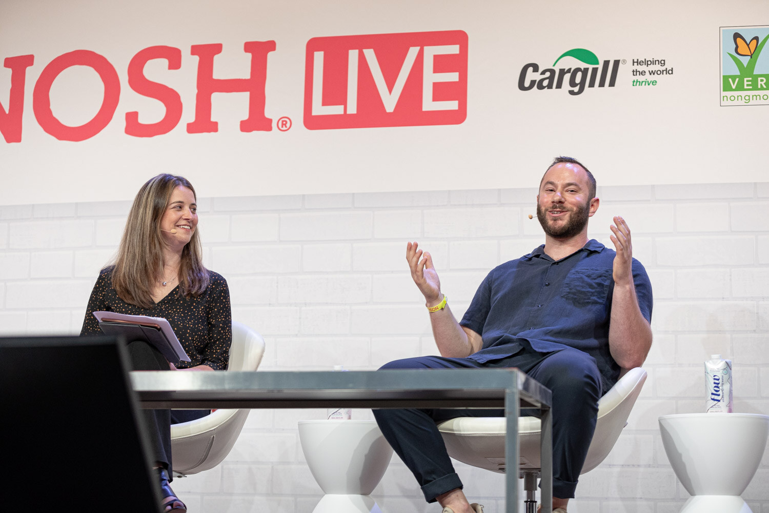 Woman with a tablet and a bearded man in a navy shirt gesturing with both hands, a Nosh Live logo behind them.