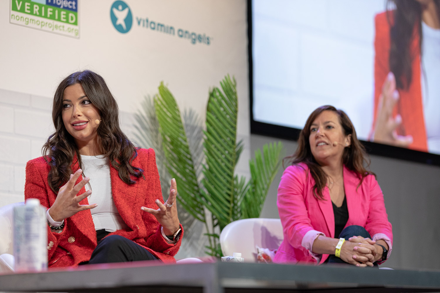 Woman in a red blazer gesturing and a woman in a pink blazer with her hands clasped, seated side by side.