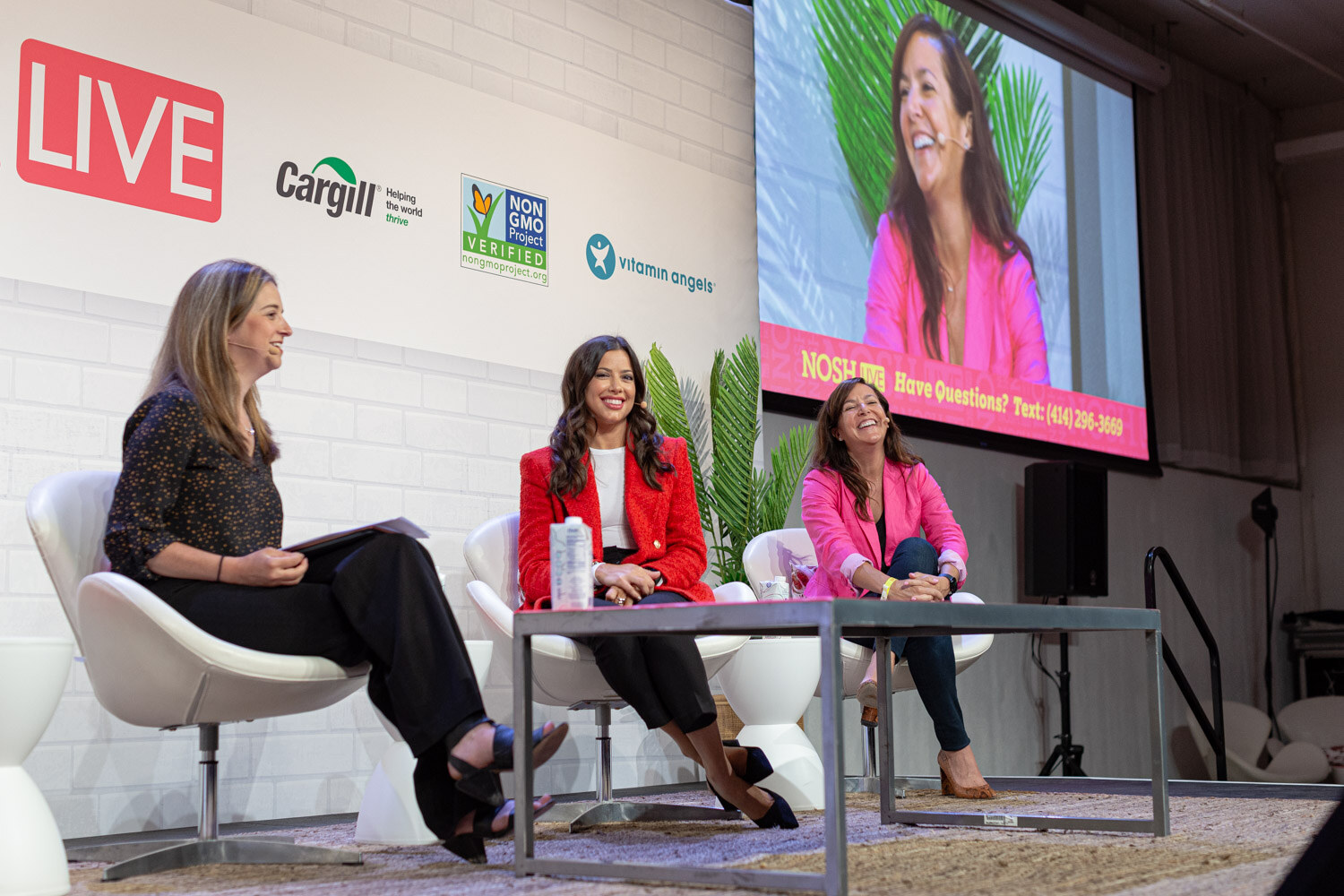 Three women in white chairs on stage, a screen at the right showing a woman in a pink blazer.