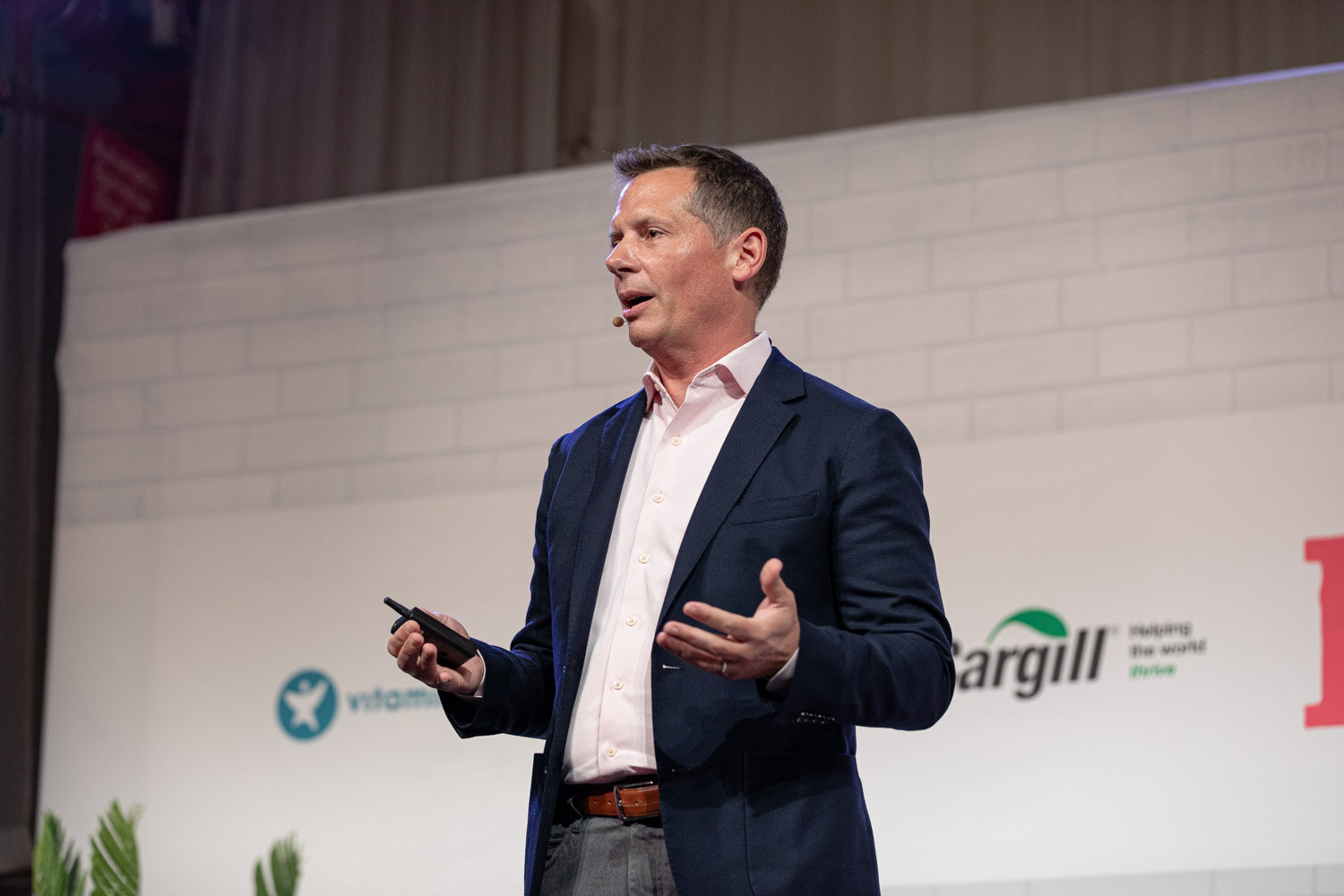 Man in a navy blazer and pink shirt with one hand open and a clicker in the other, Cargill sign behind him.