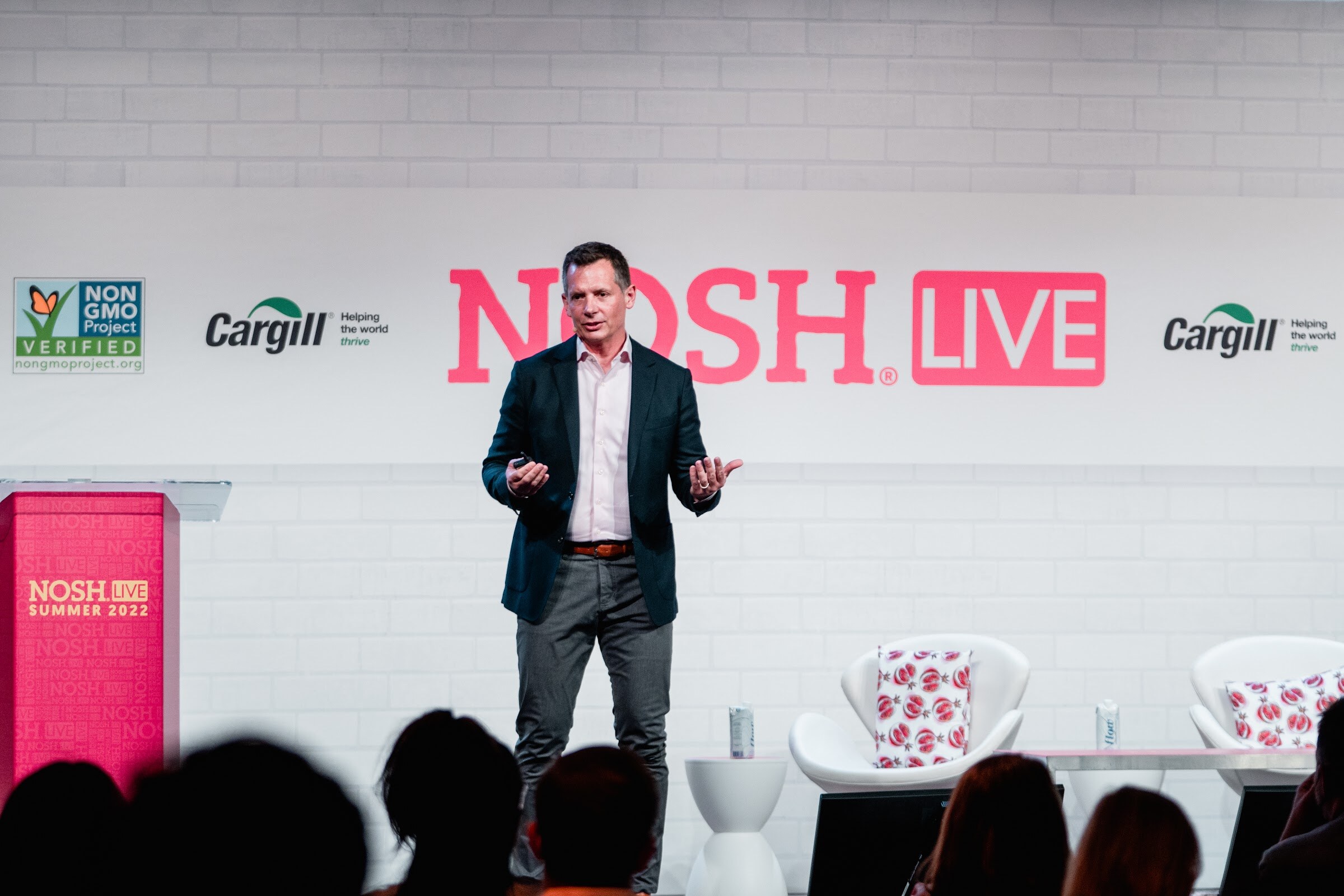 Man in a navy blazer standing near a red podium in front of a Nosh Live logo wall and two white chairs.