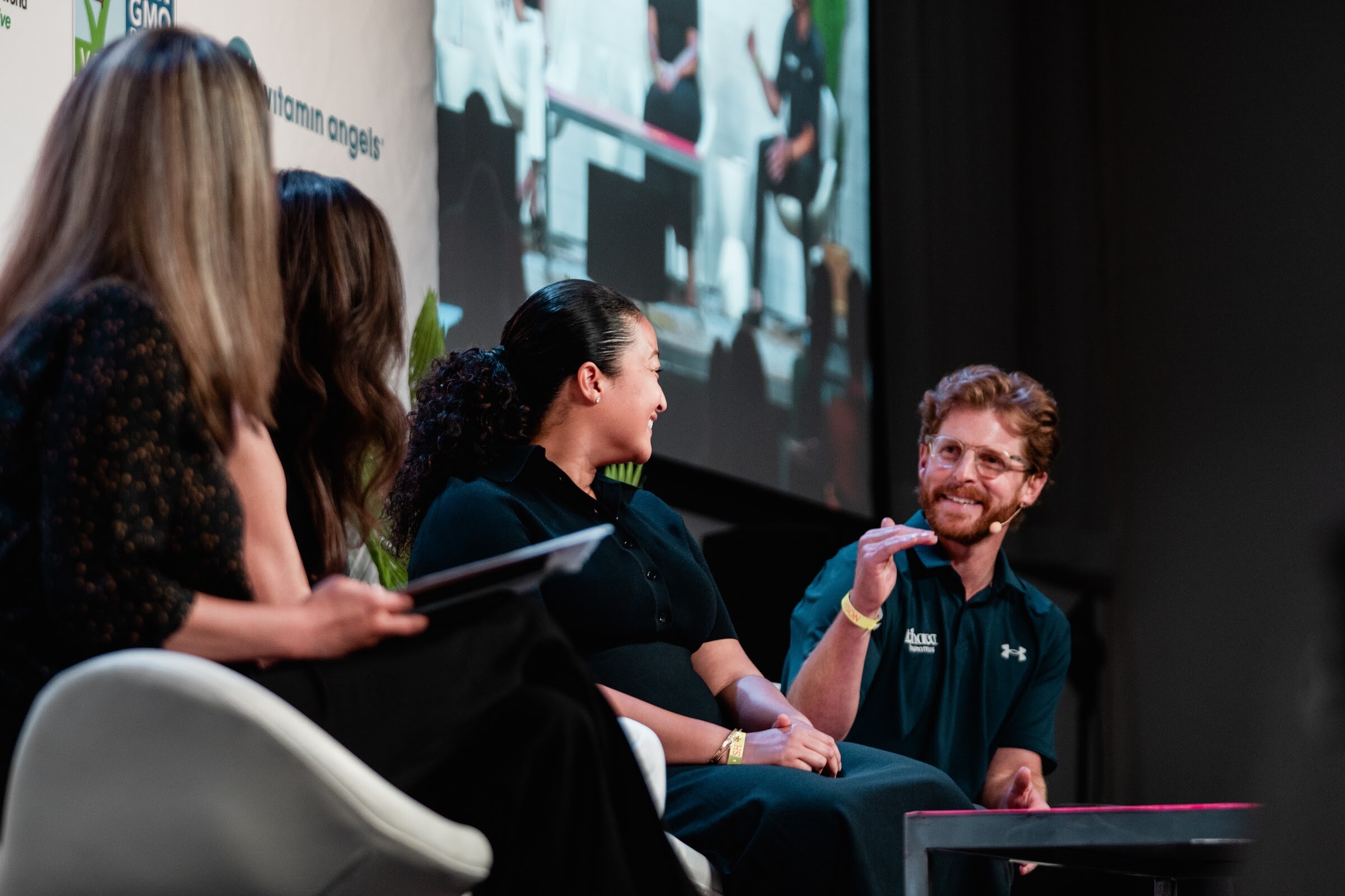 Four people seated in a row on stage, a man in a navy polo laughing as a woman in a black outfit smiles toward him.