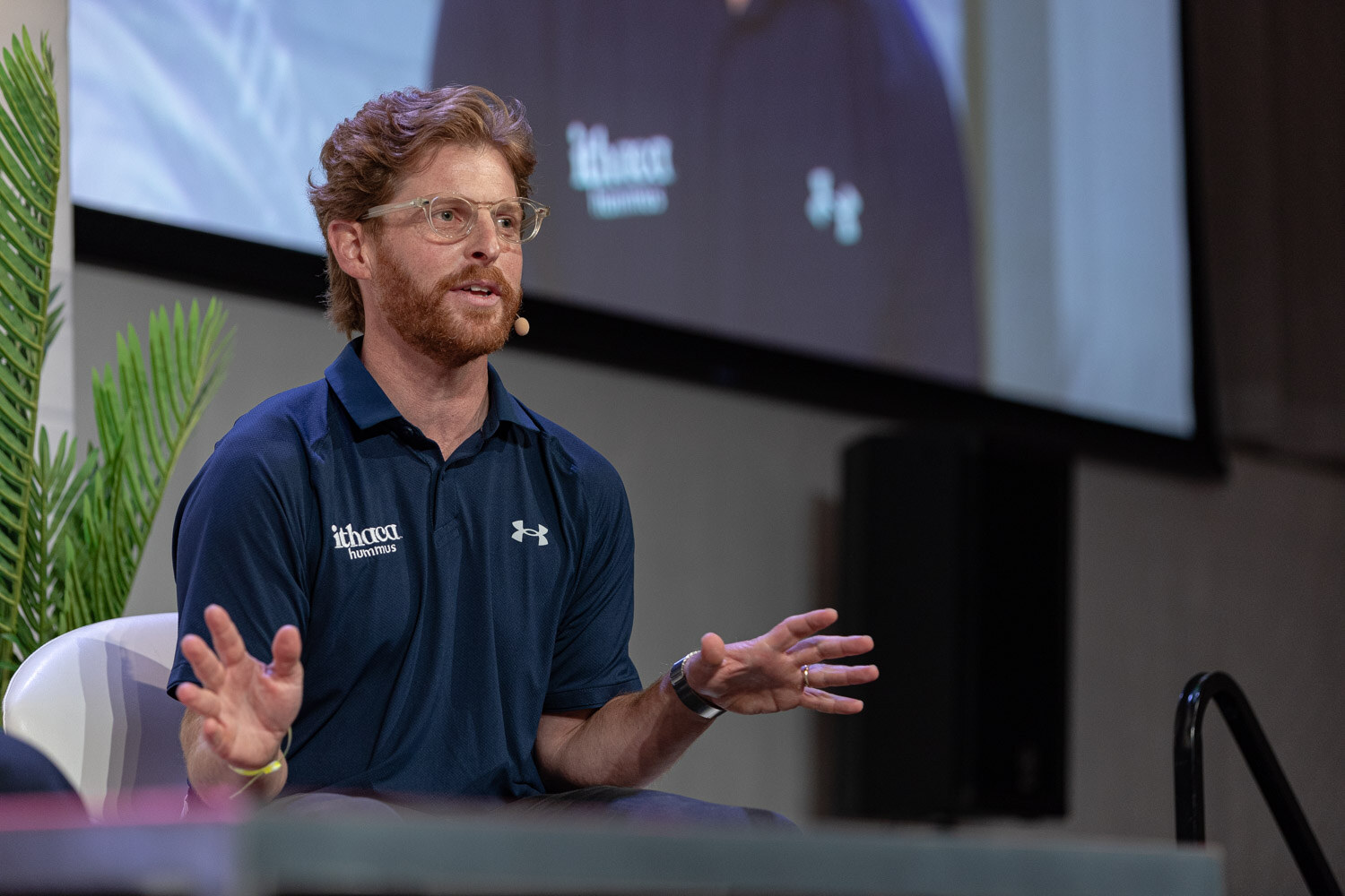 Bearded man with glasses in a navy polo shirt gesturing with both hands, a large screen behind him.