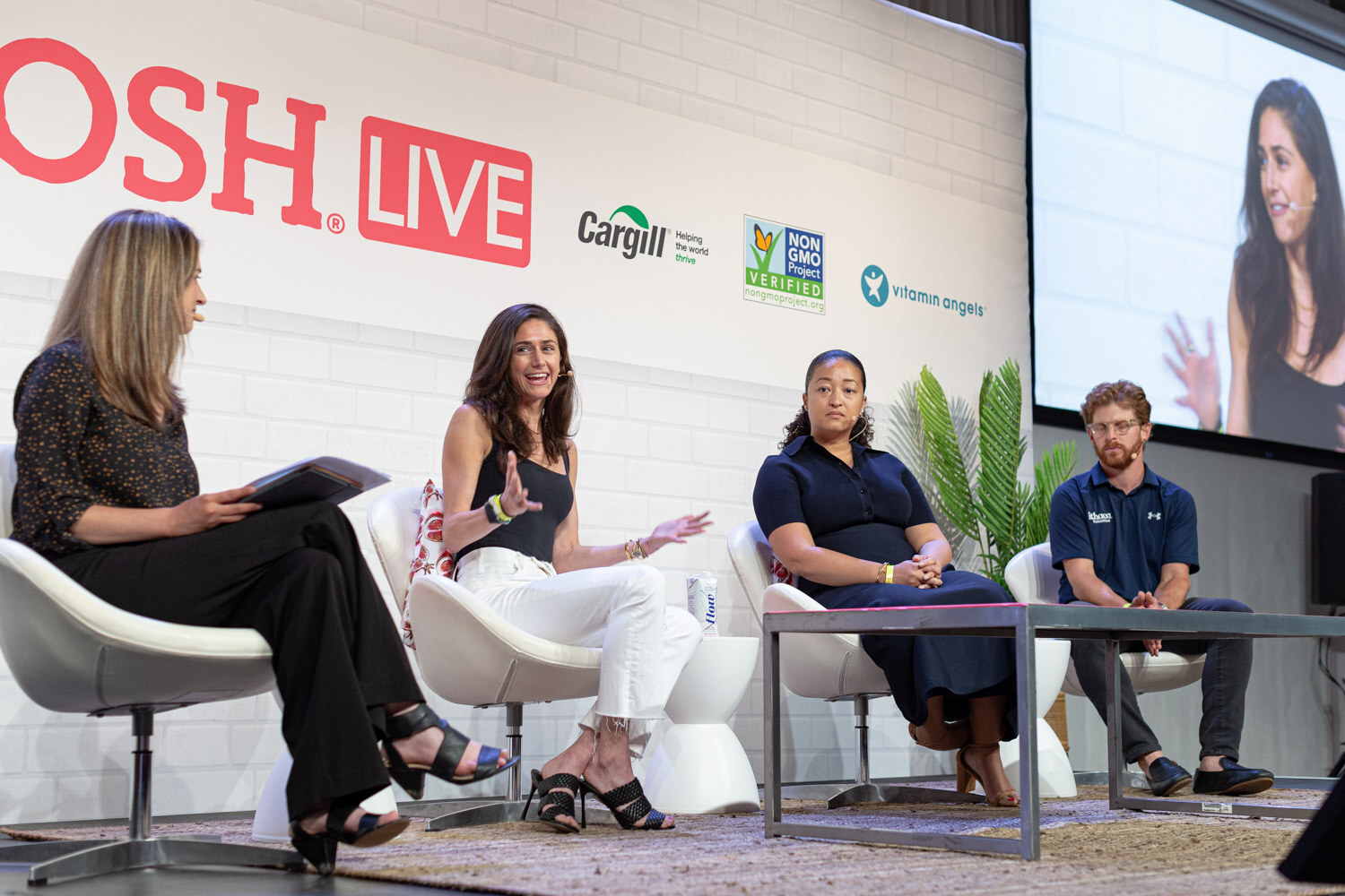 Four people seated in white chairs on stage in front of a Nosh Live wall, a screen at the right showing a close-up of one speaker.