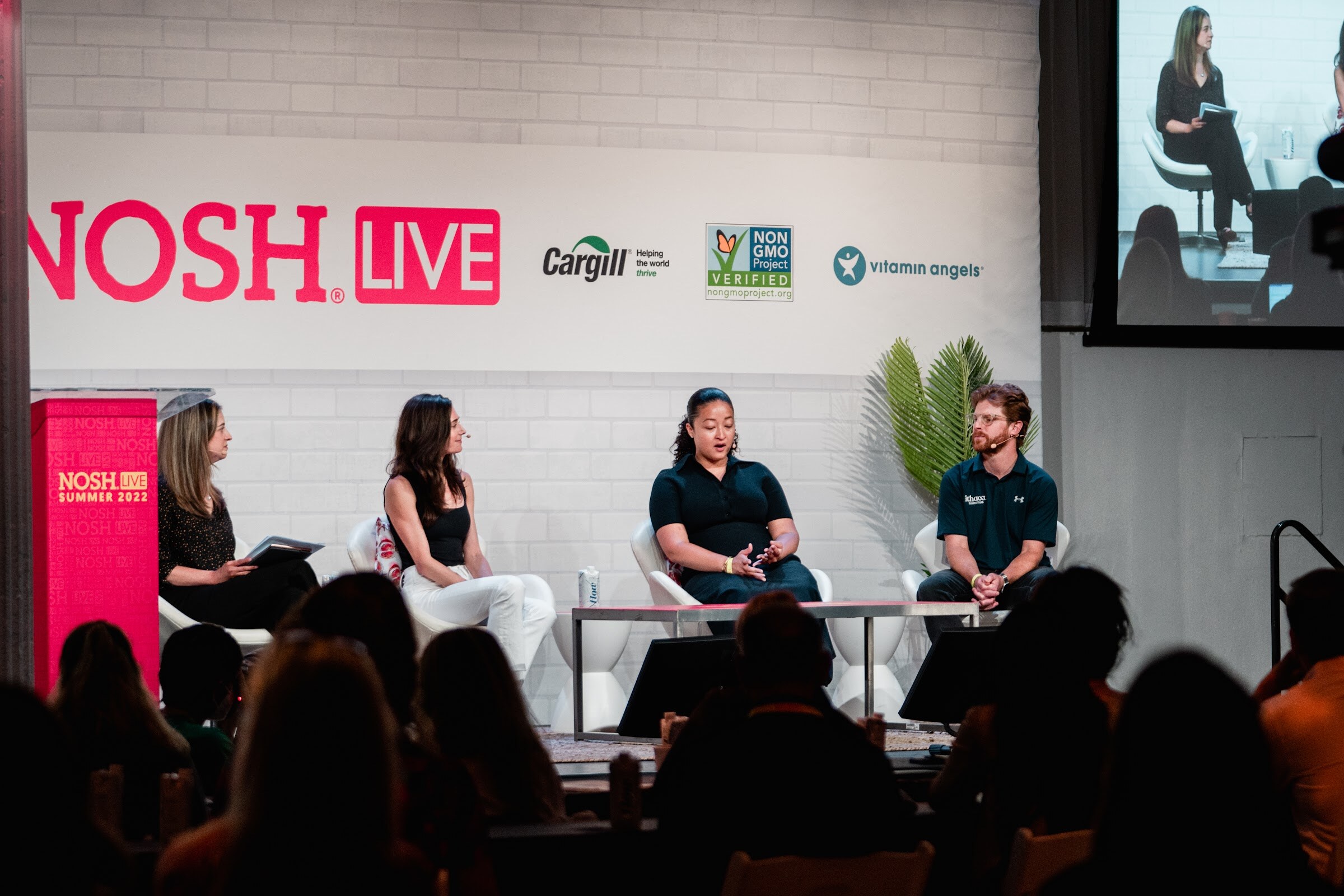 Four panelists seated on stage with audience heads in the foreground and a big screen at the right.