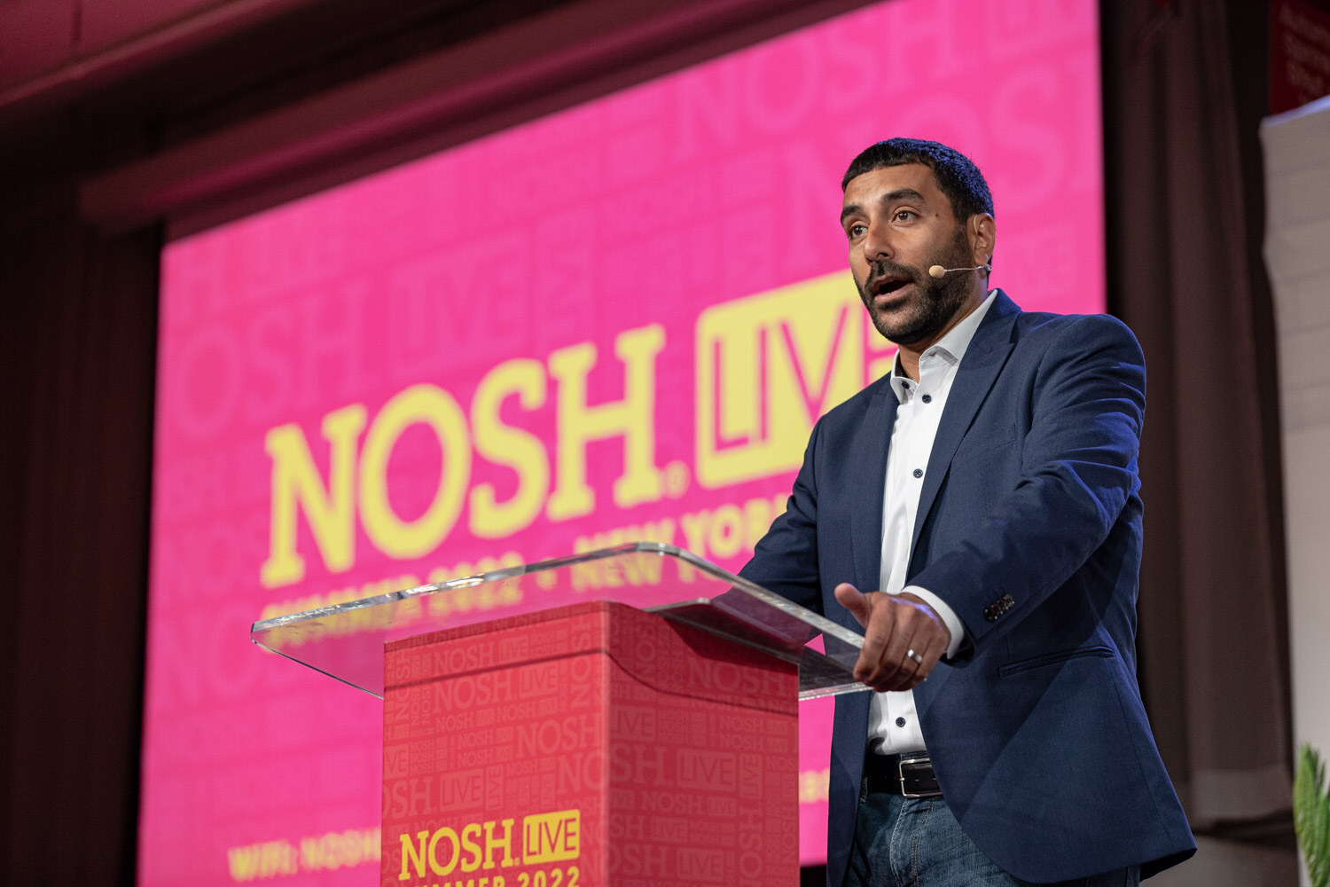 Man in a navy blazer and open collar speaking at a red podium in front of a pink Nosh Live screen.