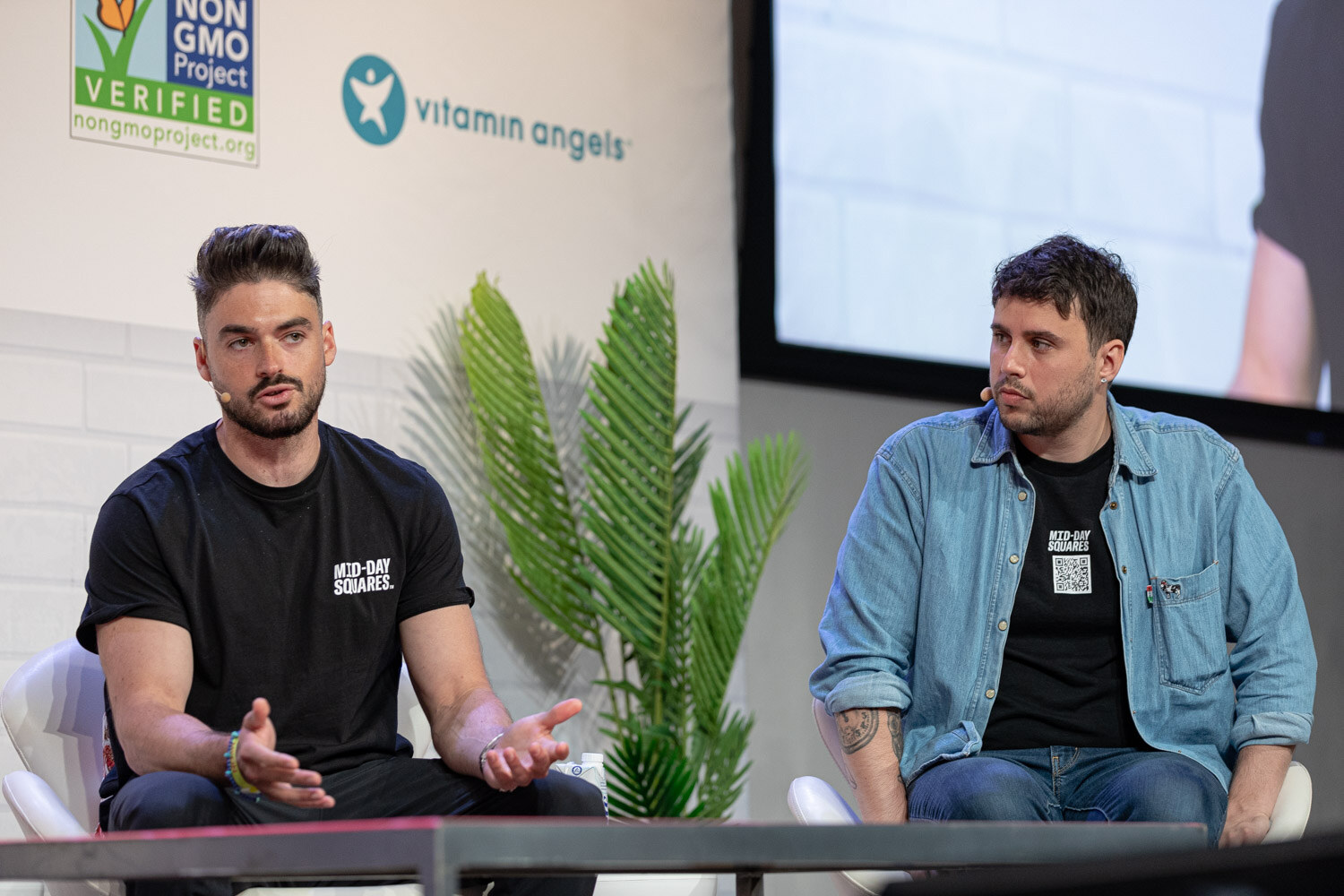 Two men seated in white chairs, one in a black Mid-Day Squares tee and one in a denim shirt, a palm plant between them.