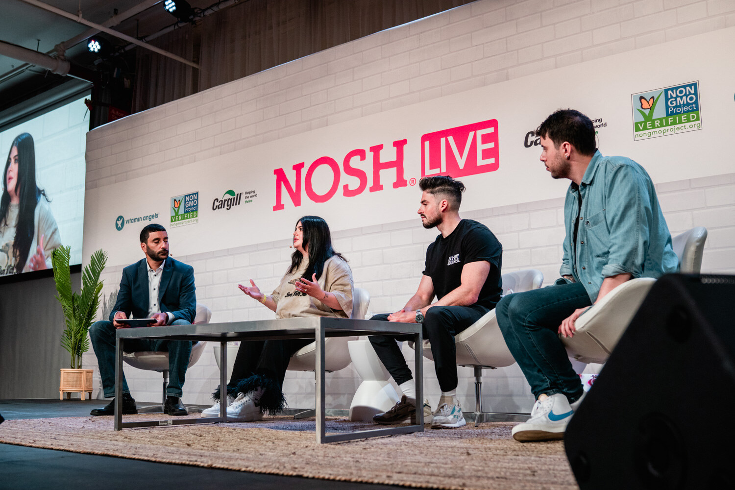 Angled view of four people seated on white chairs on a rug in front of a Nosh Live wall.