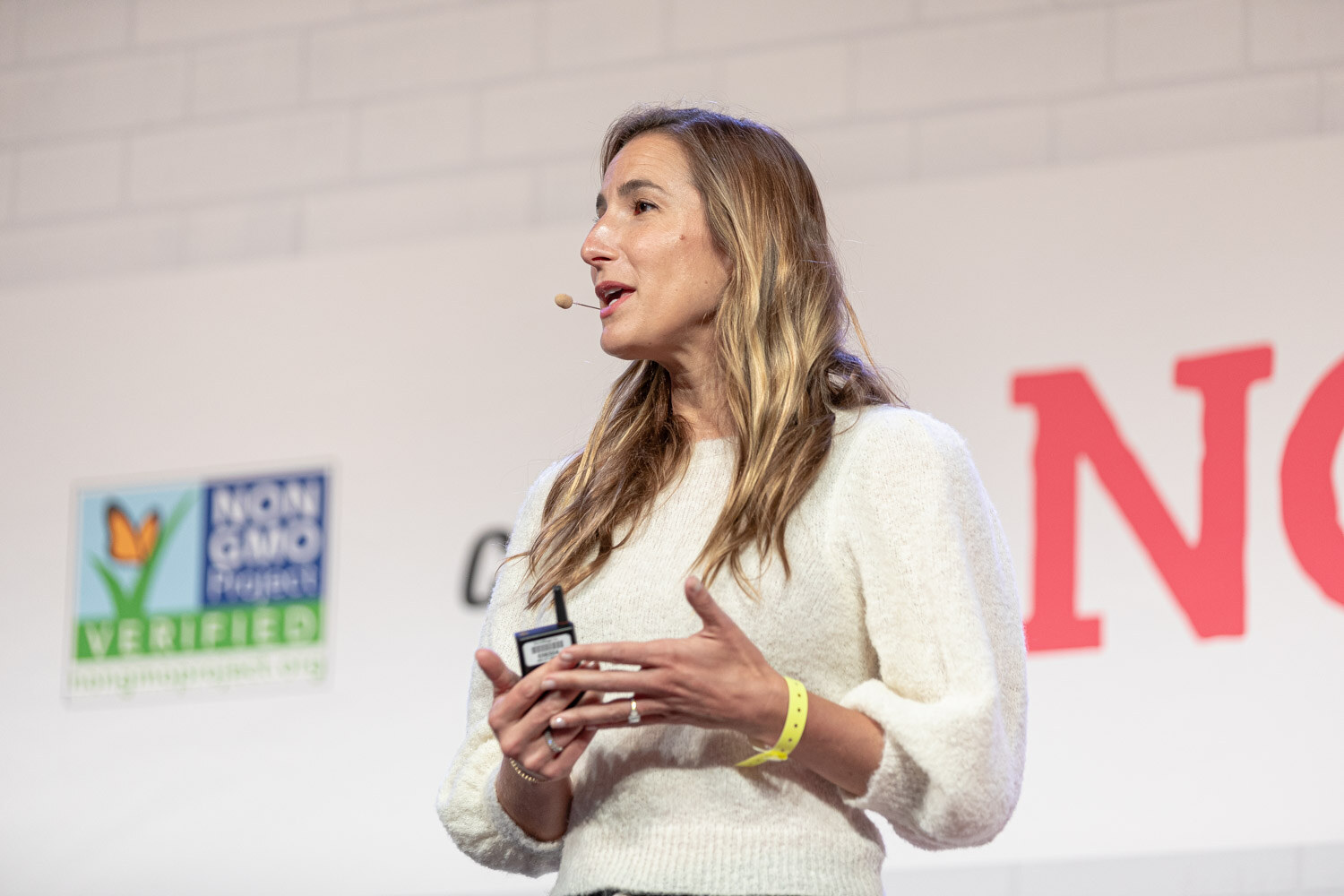 Woman with long highlighted hair in a cream sweater gesturing with both hands, a Non-GMO Project sign behind her.