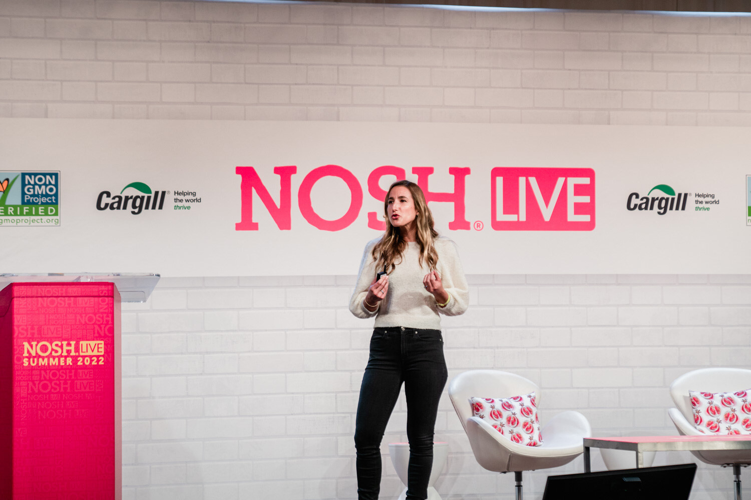 Woman in a cream sweater and black jeans standing on stage in front of the Nosh Live wall.