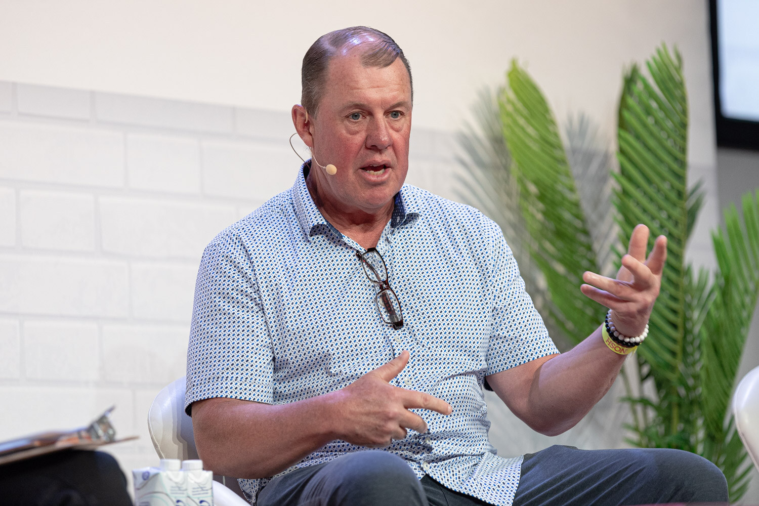 Man with a headset mic in a patterned short-sleeve shirt gesturing with both hands, palm fronds behind him.