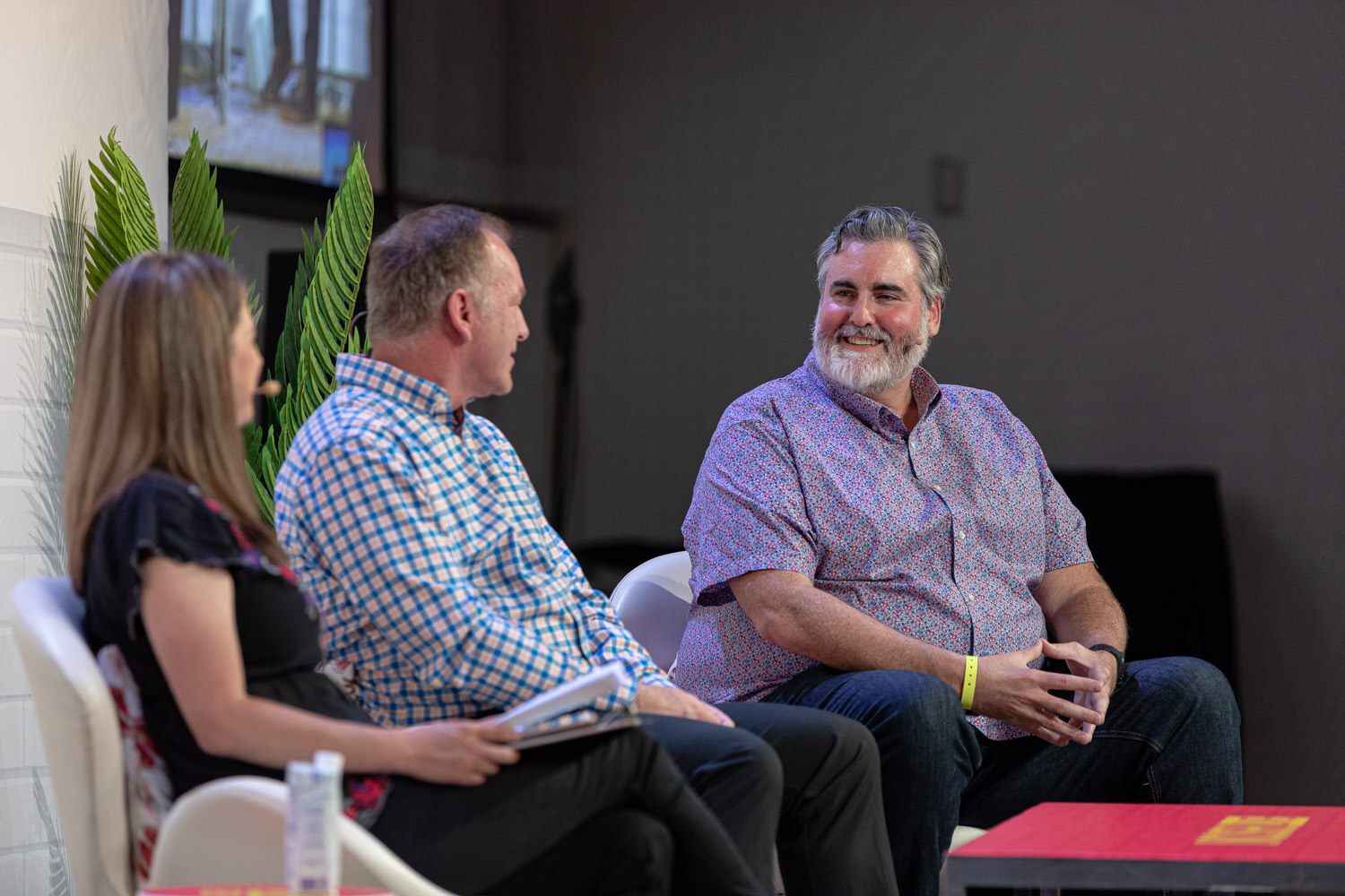 Three people seated side by side on stage, a bearded man in a purple shirt laughing at right.