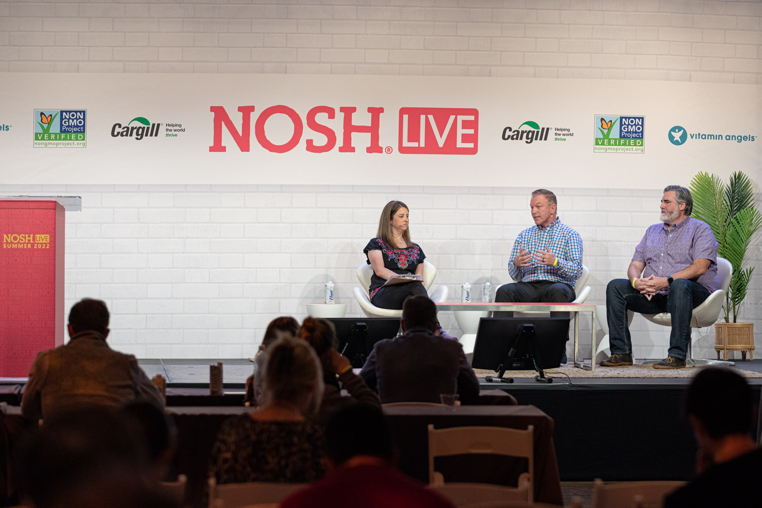 Three speakers seated on stage under the Nosh Live logo with audience heads and a red podium in the frame.