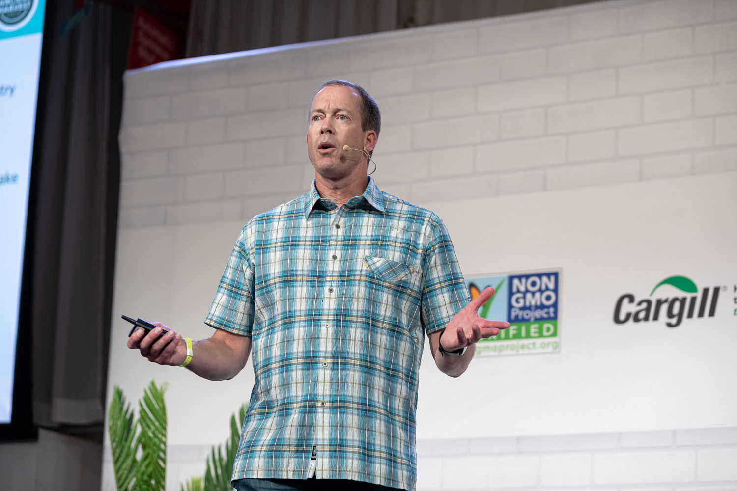 Man with a headset mic in a teal plaid shirt holding a clicker, a Non-GMO Project sign behind him.