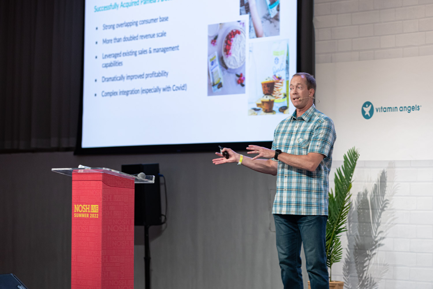 Man in a teal plaid shirt standing beside a red podium, a screen showing bullet points and photos of baked goods.