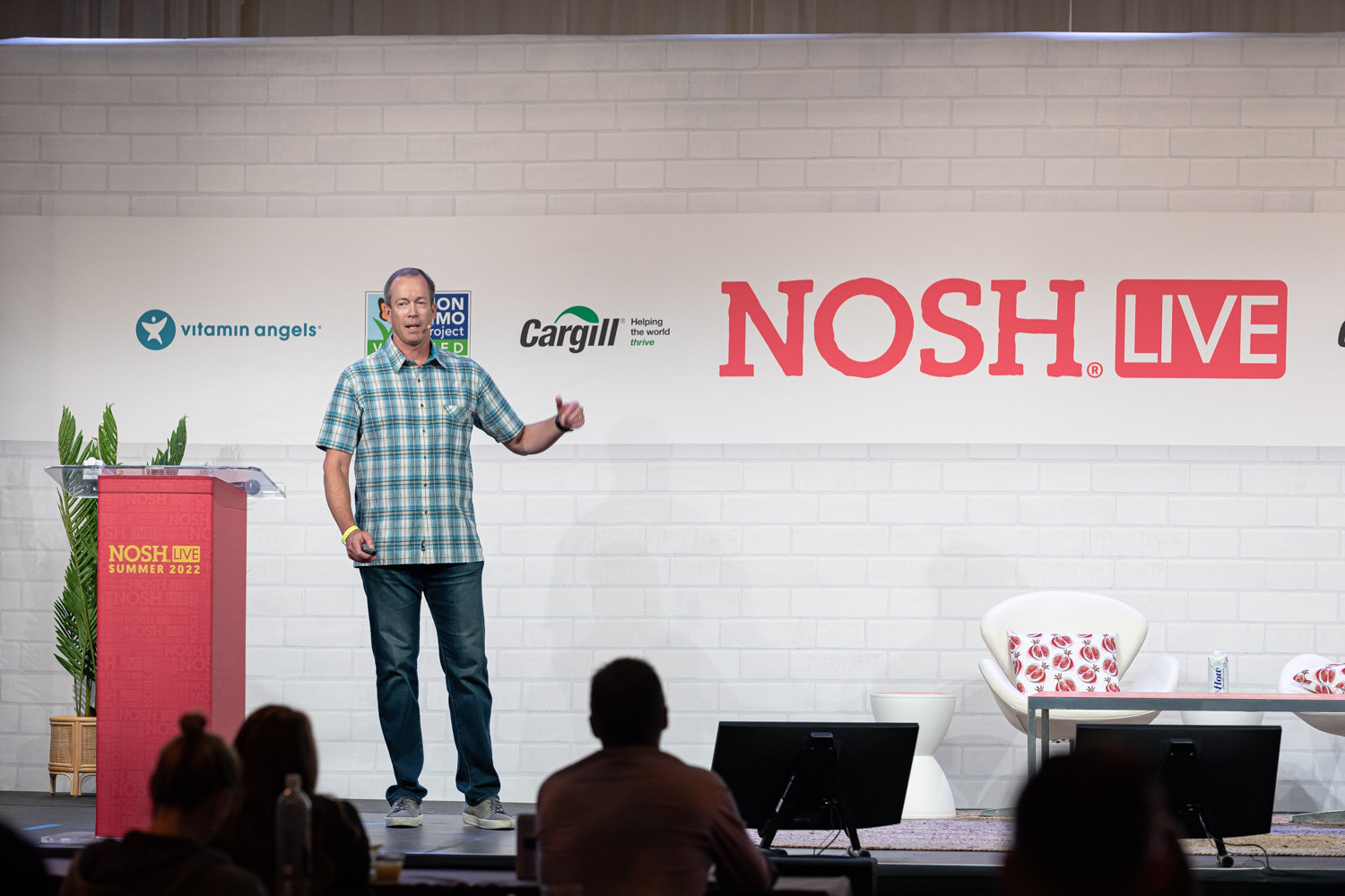 Man in a plaid shirt standing near a red podium and white chairs, audience silhouettes in front.