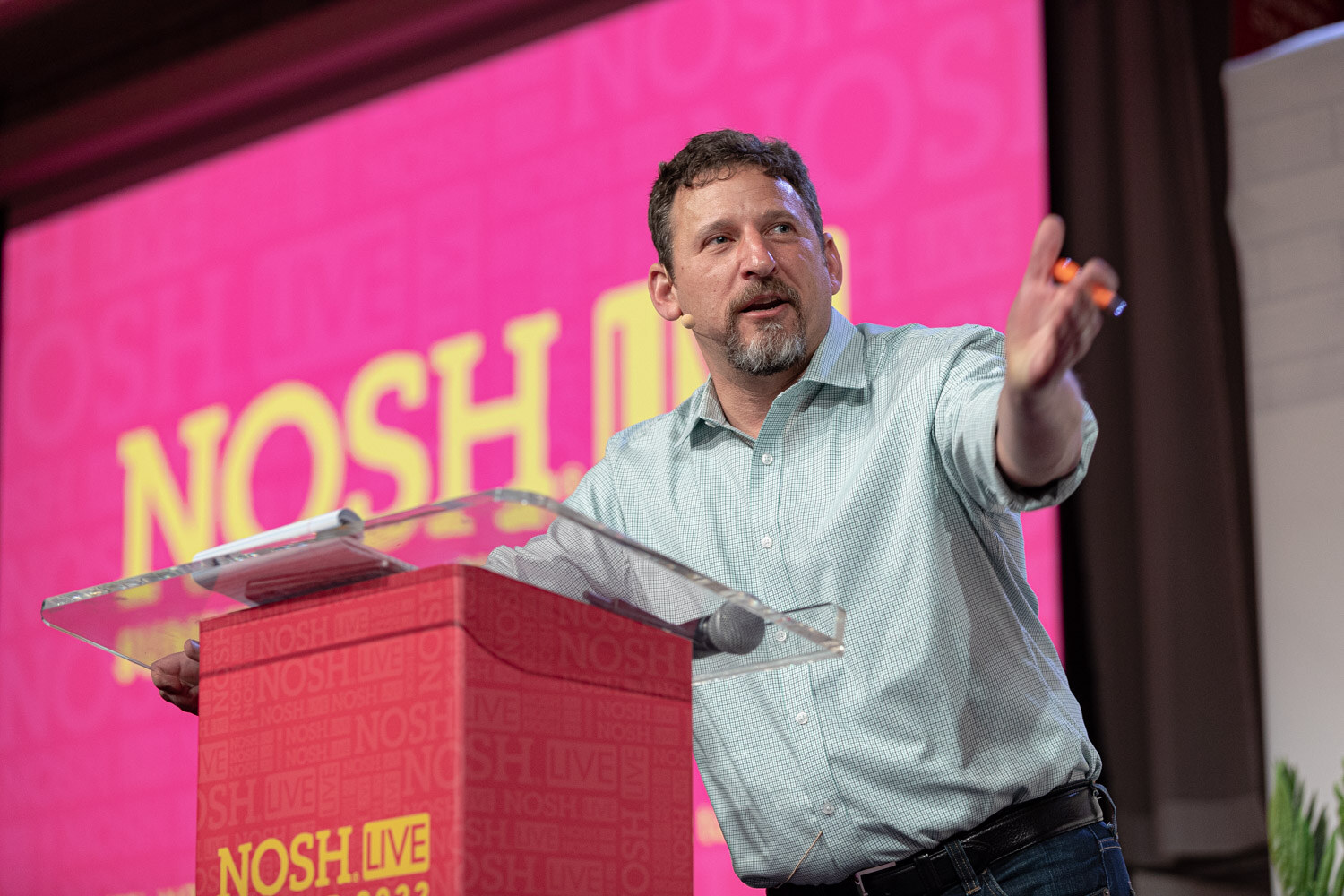 Man with a gray beard in a pale green shirt leaning over a clear podium, one arm outstretched, a pink Nosh Live screen behind him.