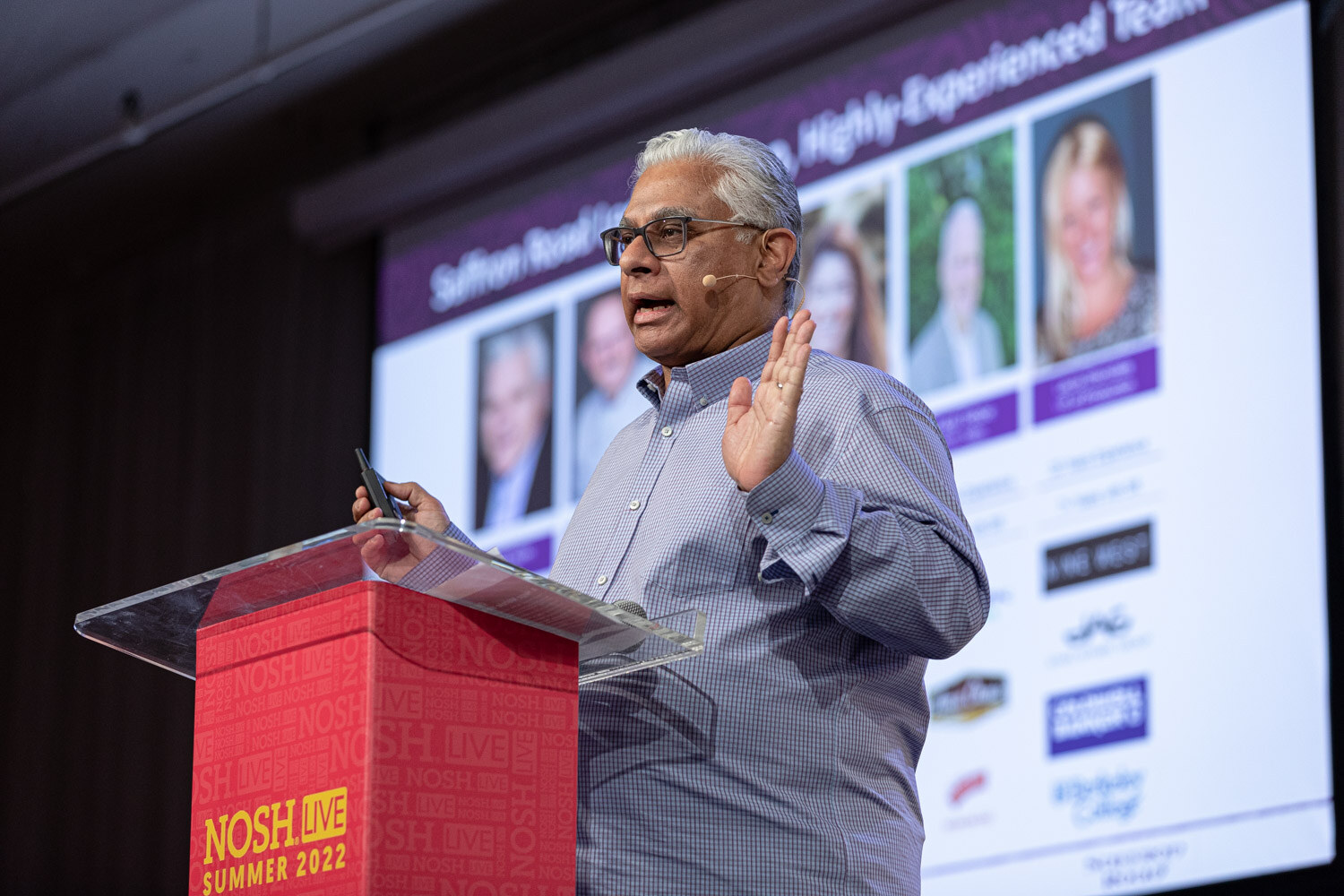 Gray-haired man in glasses and a blue shirt with one hand raised at a podium, a slide with team photos behind him.