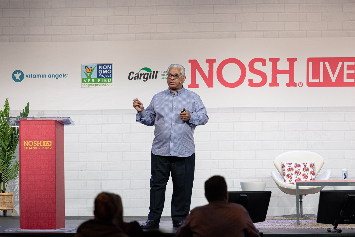 Gray-haired man in a blue shirt gesturing near a red podium in front of the Nosh Live wall, audience heads in the foreground.