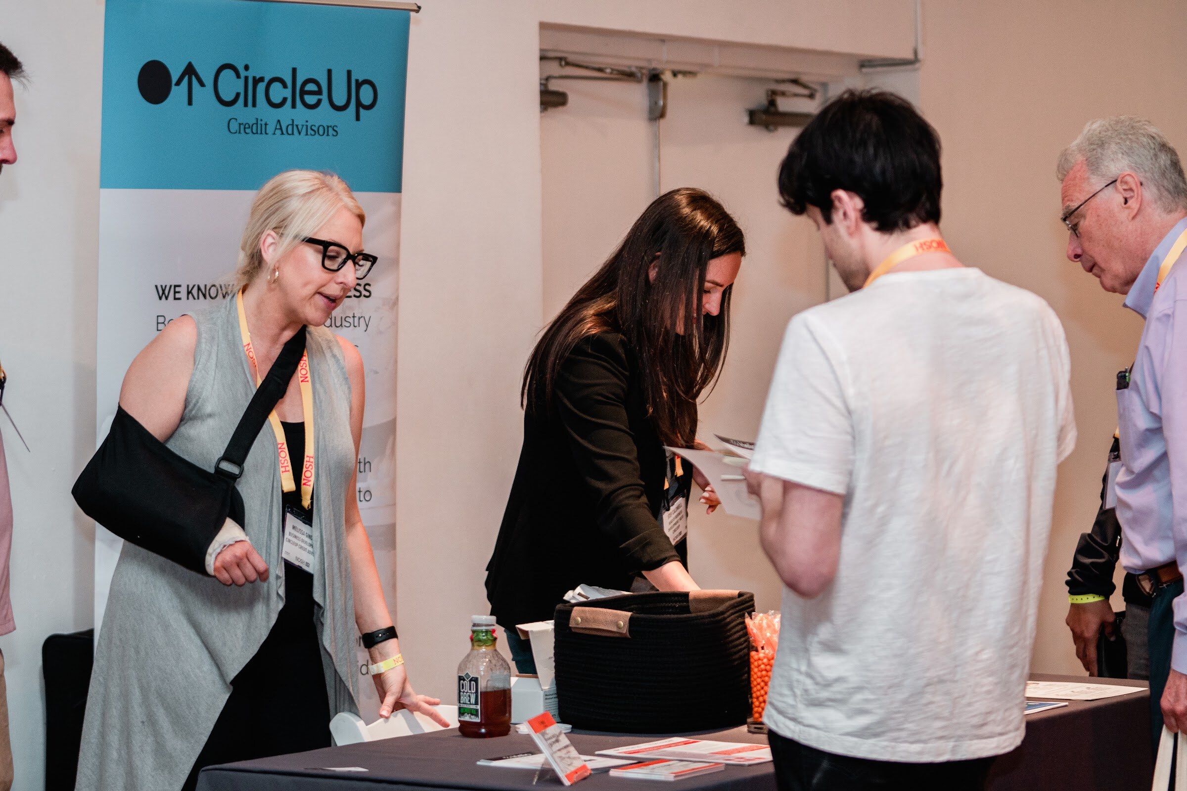 Woman with her arm in a sling and a woman in black greet attendees at a dark table beside a CircleUp Credit Advisors banner.
