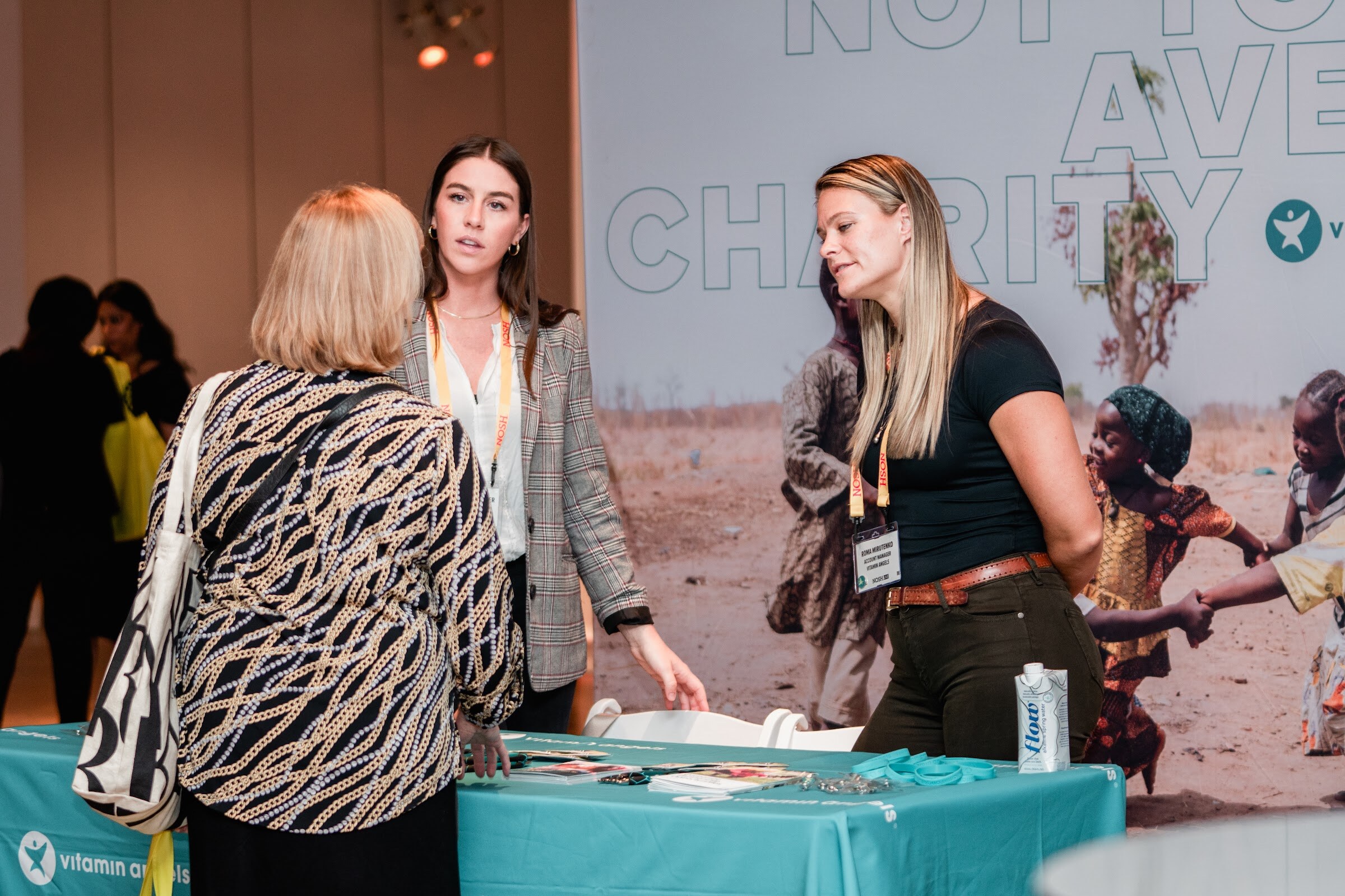 Two women with lanyards chat with an attendee in front of a Not Your Average Charity backdrop and a teal Vitamin Angels tablecloth.