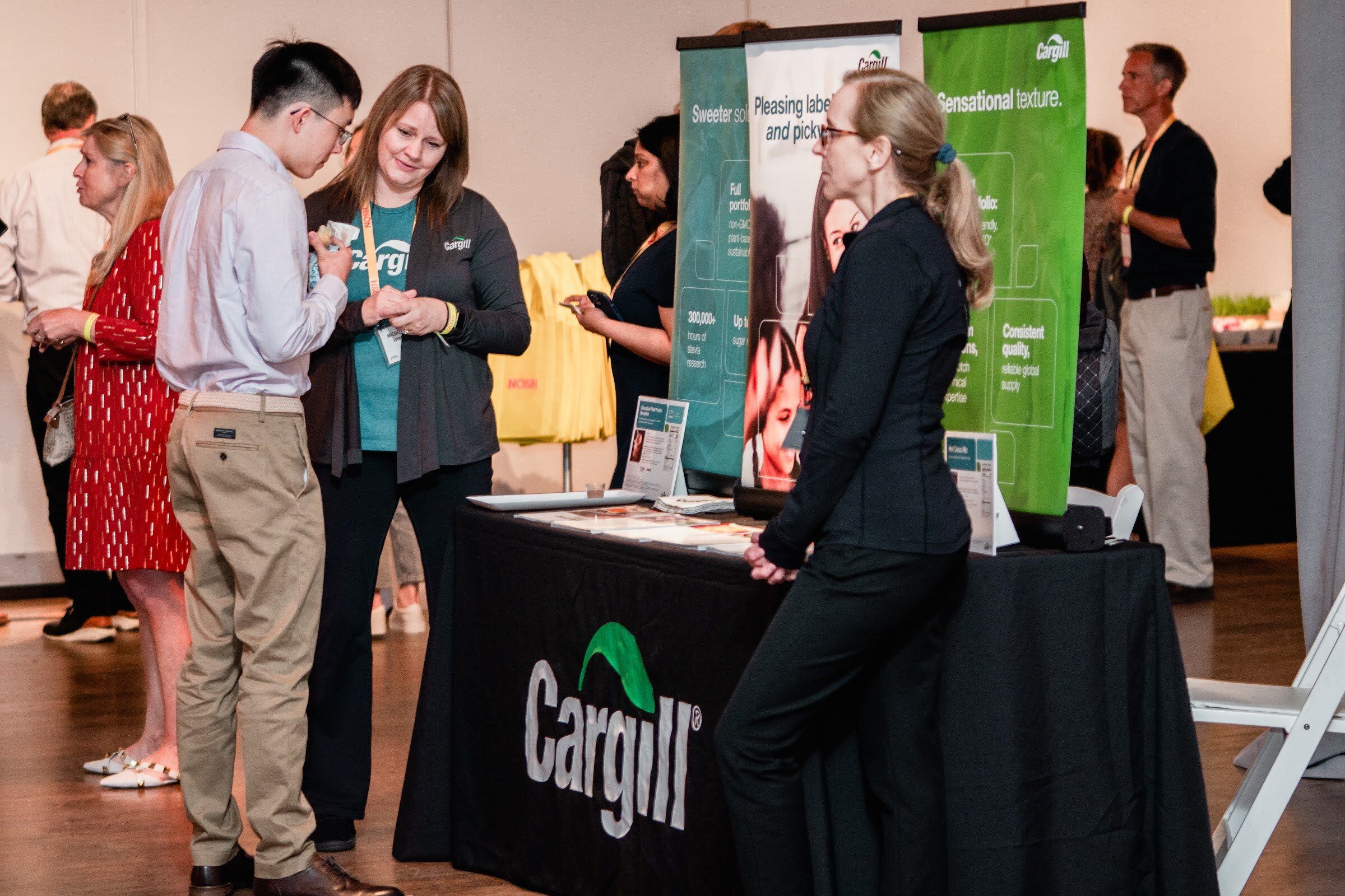 Group of attendees talking beside a black Cargill table and green and teal Cargill banners reading Pleasing label-readers and picky eaters.