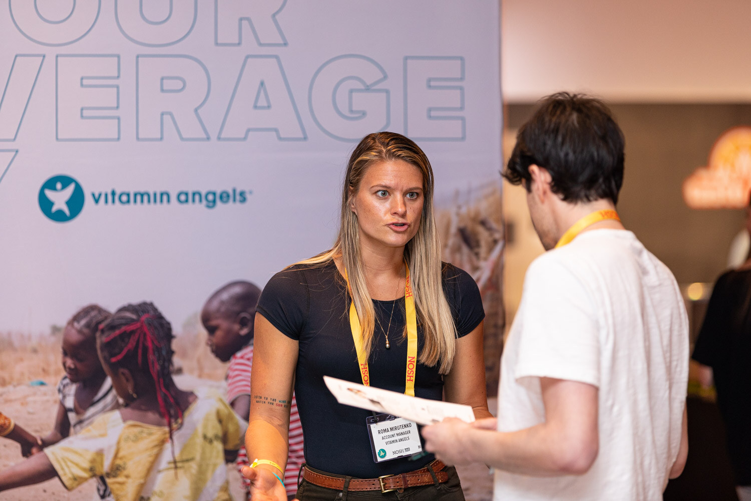 Blond woman with a lanyard holding a brochure while a man in a white tee listens, in front of a Vitamin Angels backdrop.