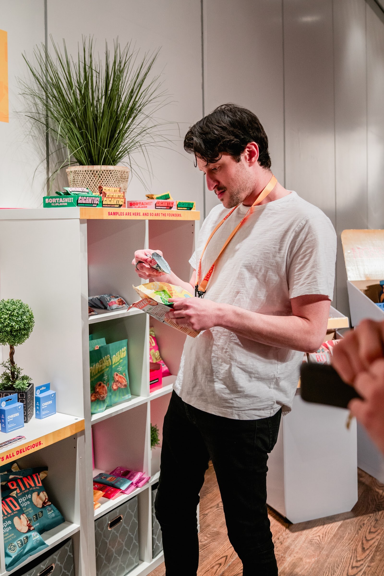 Man in a white tee with an orange lanyard holding a snack package beside white shelves and a potted plant.