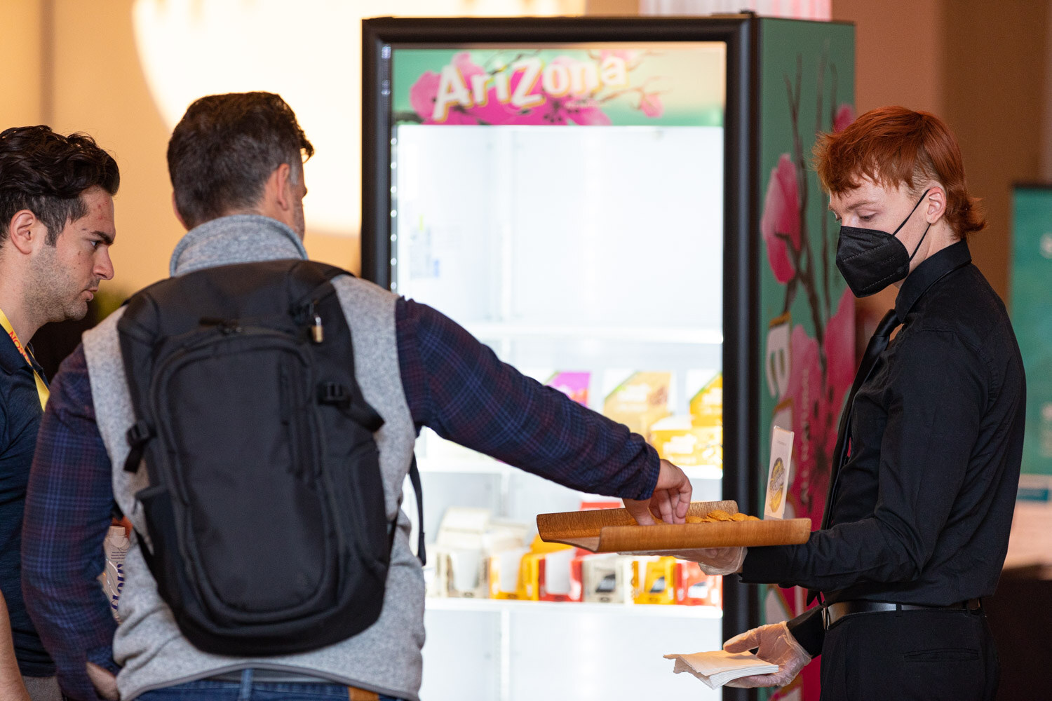 Man with a backpack and a masked server holding a wooden tray next to a glass-door cooler with an Arizona design.