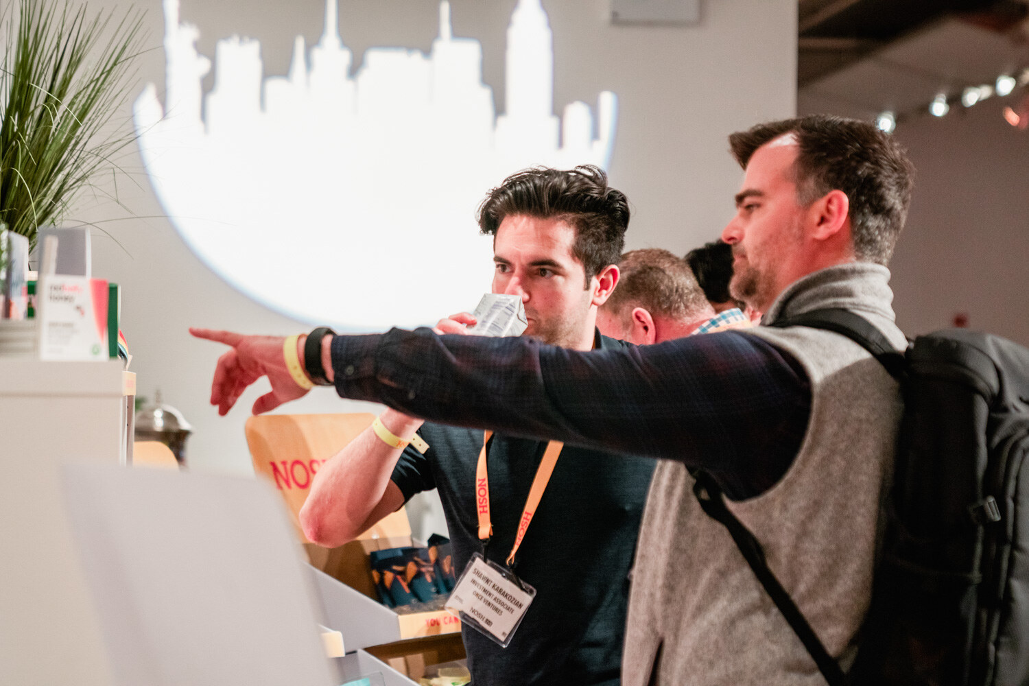 Man in a navy tee with a lanyard tasting a sample beside a man pointing, a white New York skyline projection behind them.