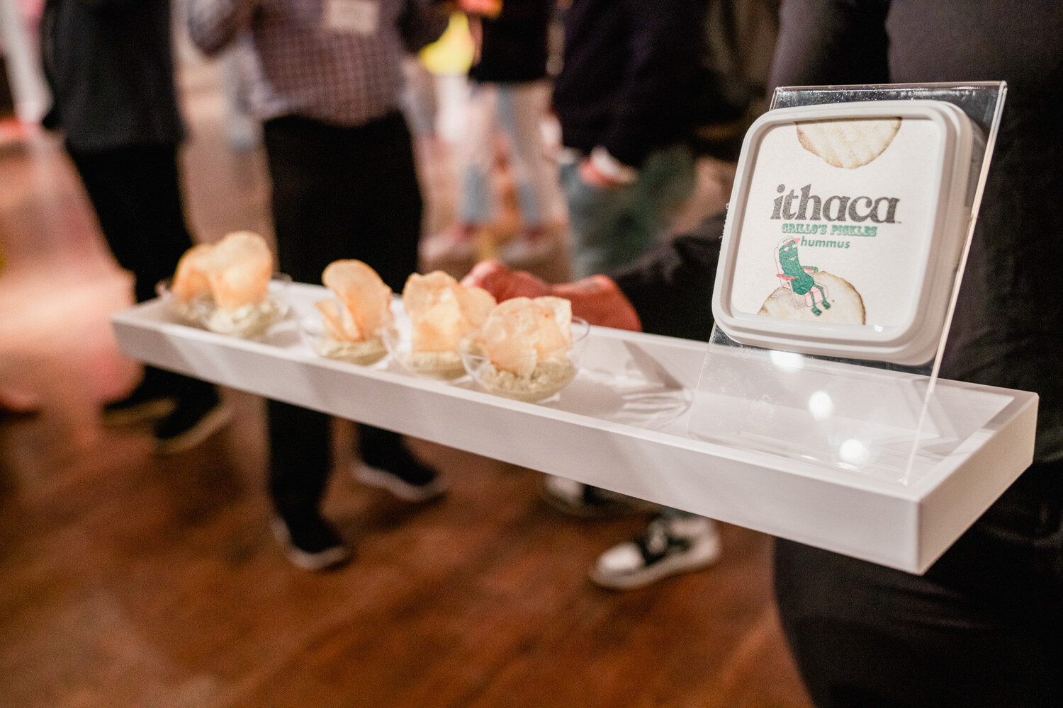 White serving tray with five samples in cups and an Ithaca hummus container, attendees blurred in the background.