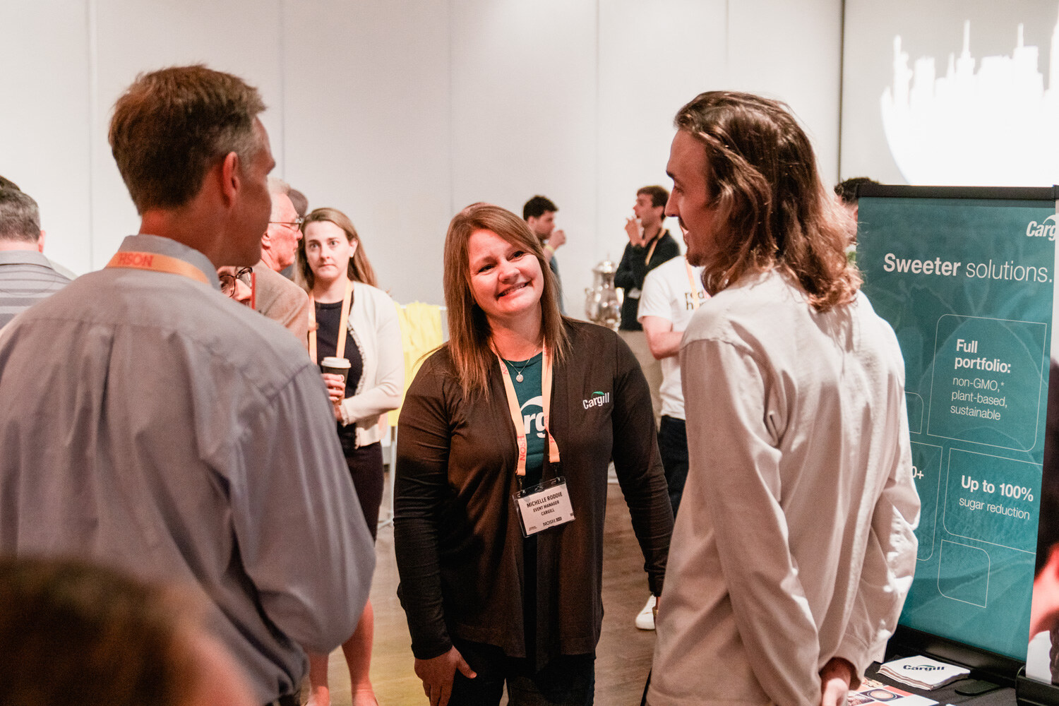 Woman in a dark Cargill shirt with a lanyard talking with a man in a gray shirt and a man with long hair near a Sweeter solutions banner.