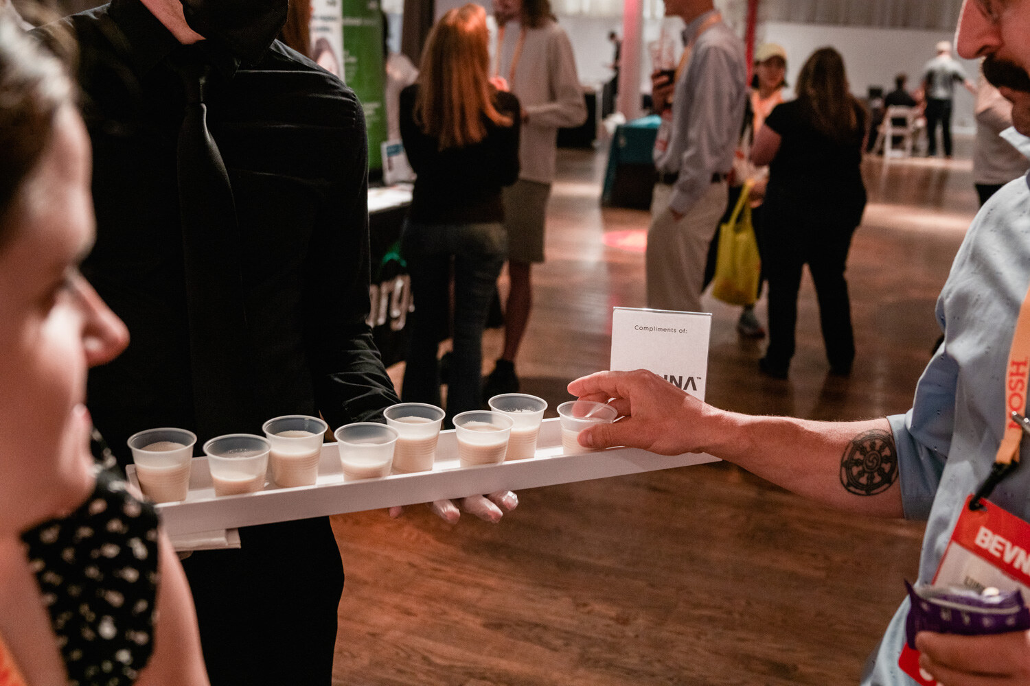 Tray holding rows of small cups of white drink with attendees in the background.