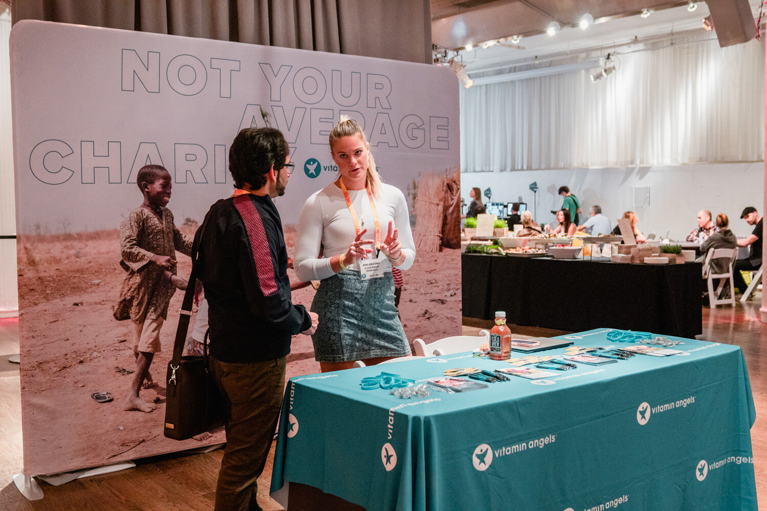 Woman in a white top and denim skirt beside a man in a dark jacket in front of a Not Your Average Charity backdrop and a teal table.