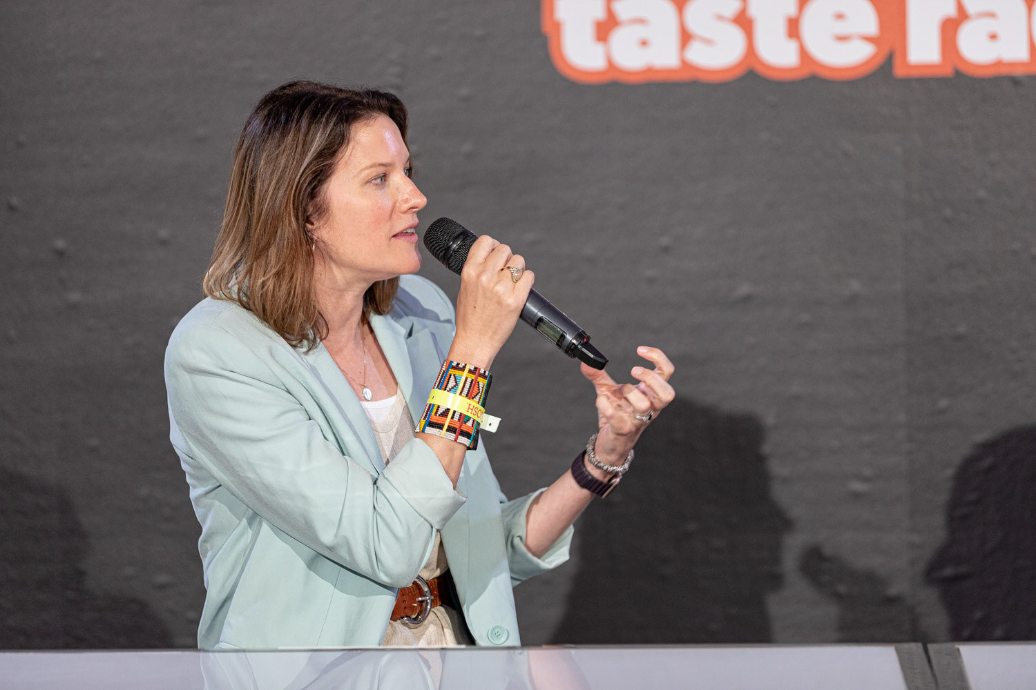 Woman in a mint-green blazer and stacked wristbands holding a mic in front of a dark Taste Radio wall.