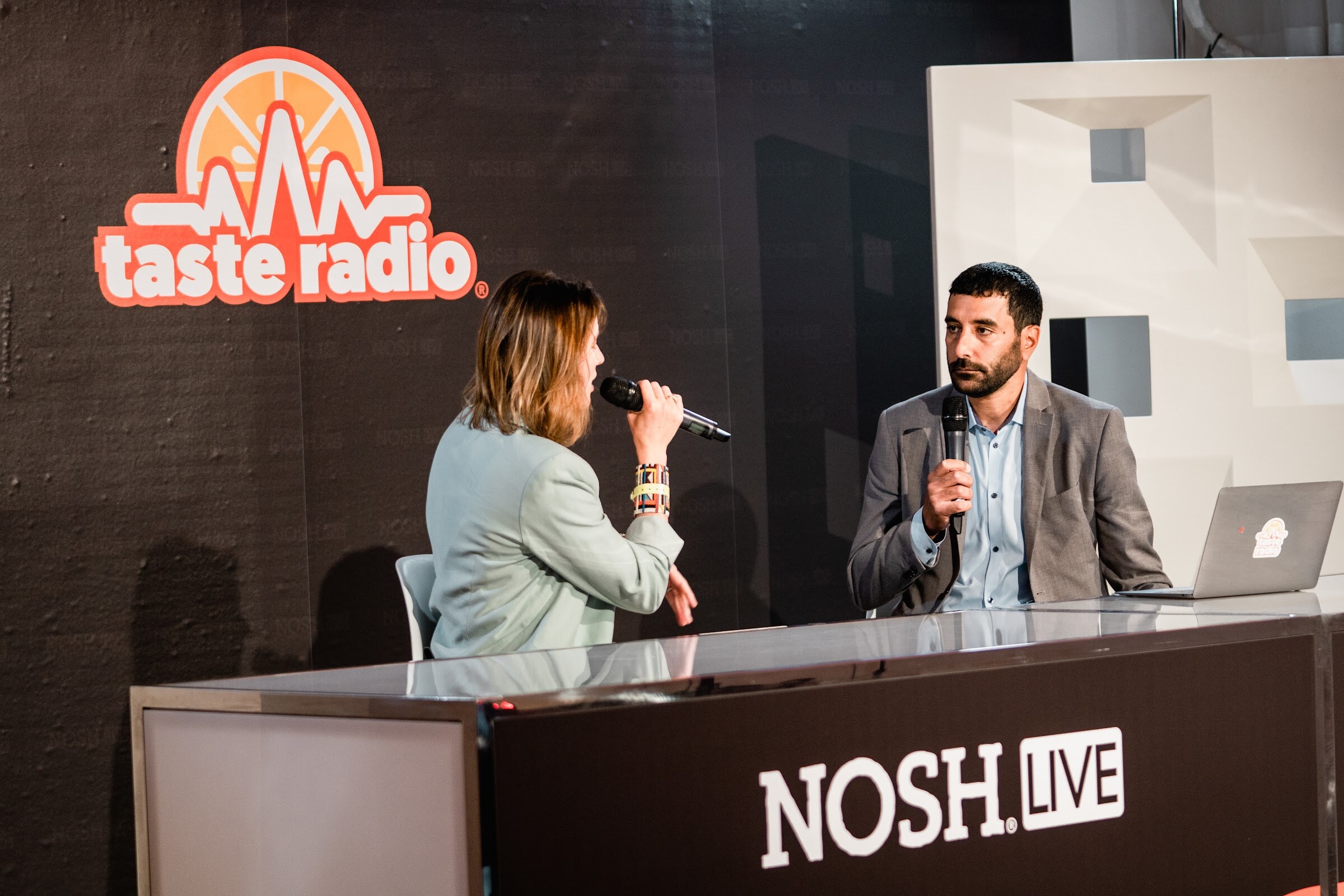 Woman in a mint blazer and man in a gray blazer holding mics at a desk marked Nosh Live in front of the Taste Radio logo.