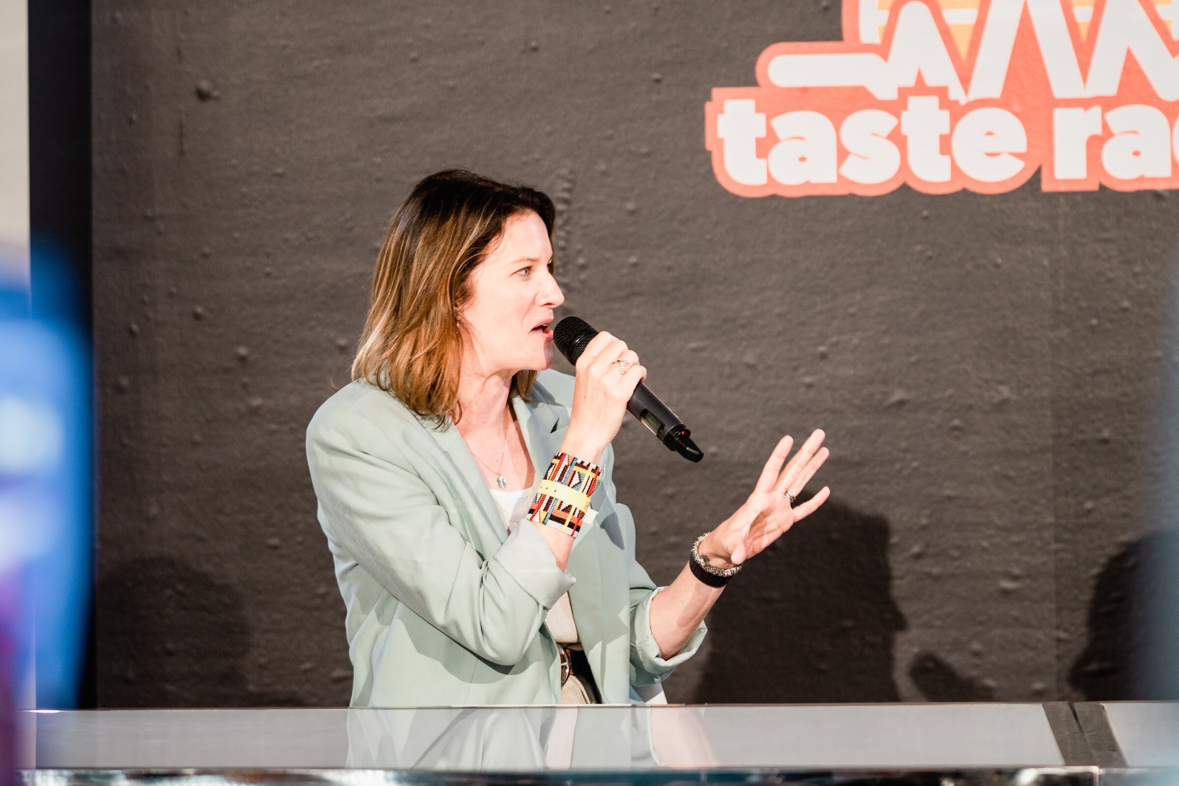 Woman in a mint blazer with a mic and open hands in front of the Taste Radio logo.