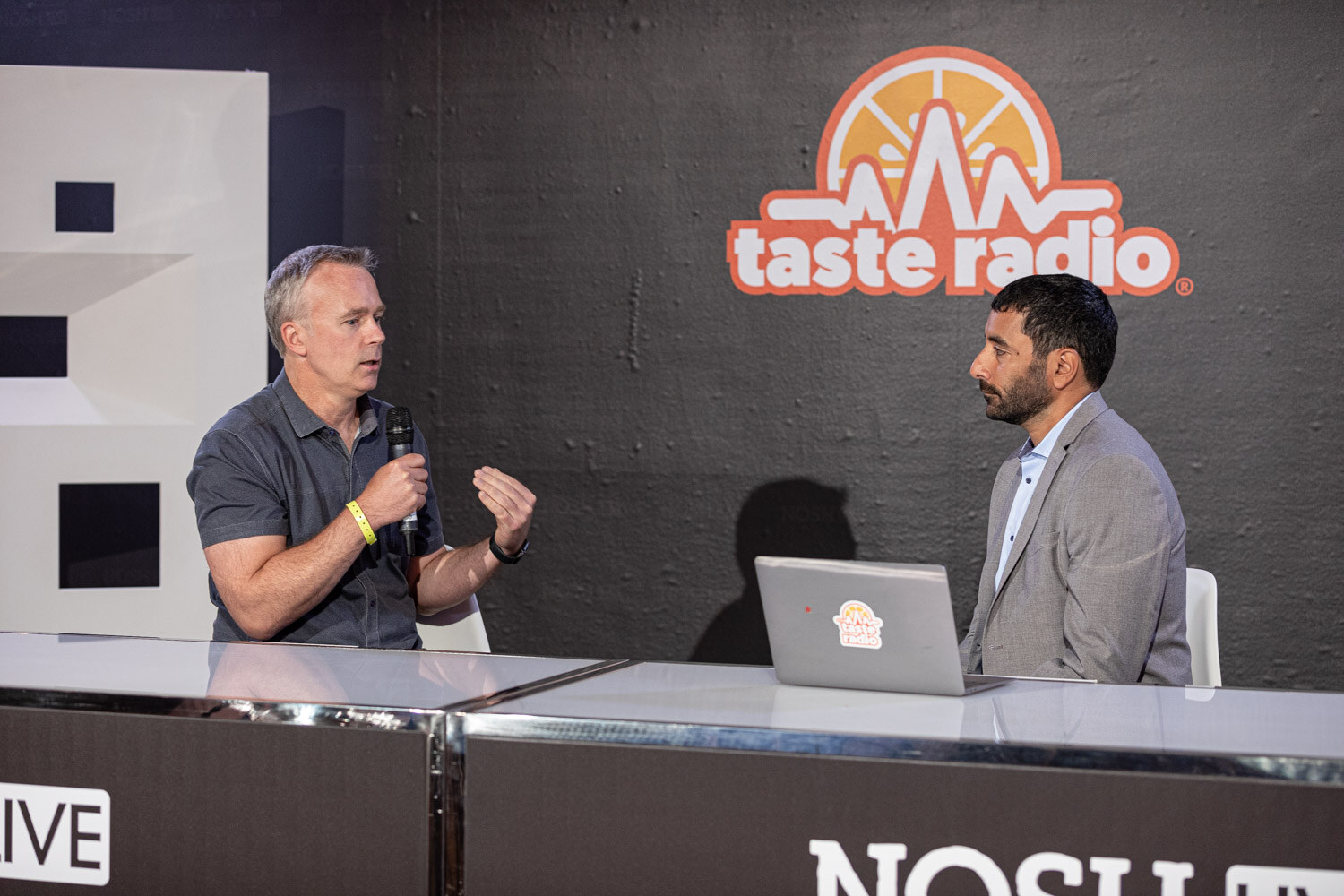 Man in a gray polo and man in a gray blazer seated at a desk with a laptop in front of the Taste Radio logo.
