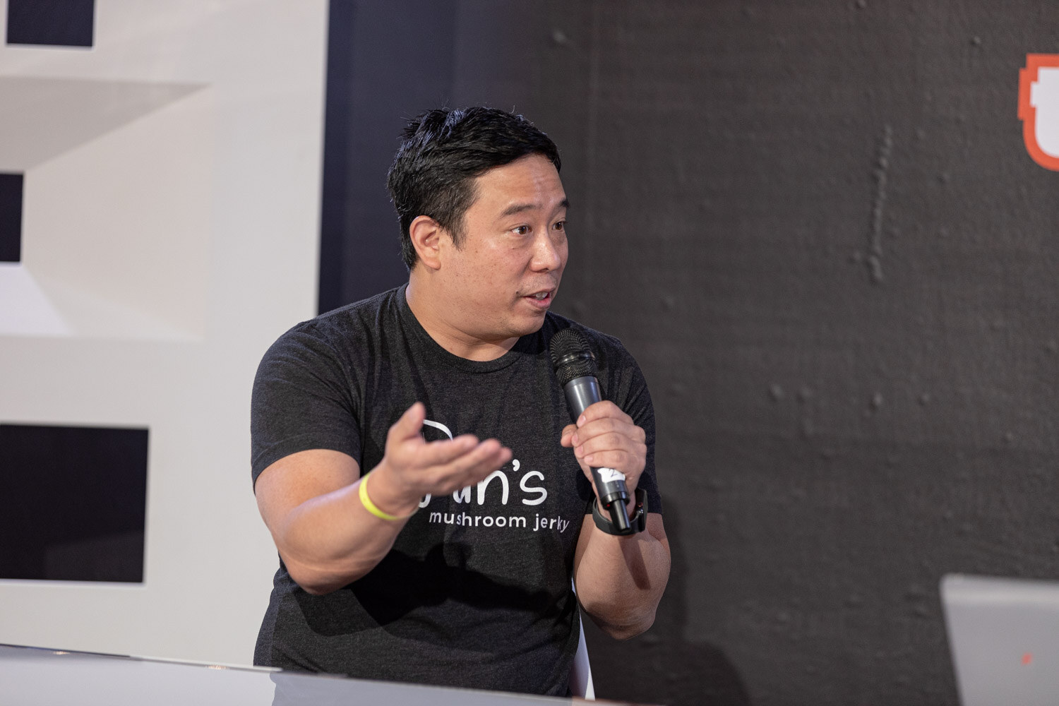 Man in a black Pan's mushroom jerky t-shirt with a mic, one hand open.