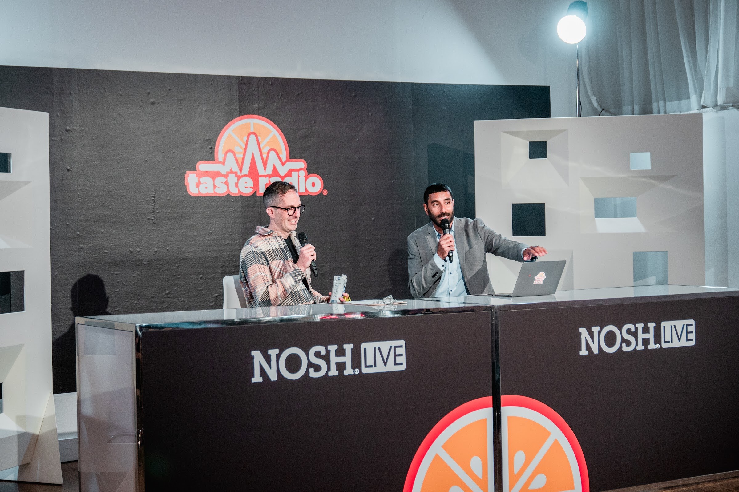 Two people seated at a long desk branded Nosh Live in front of a Taste Radio sign and white set pieces.
