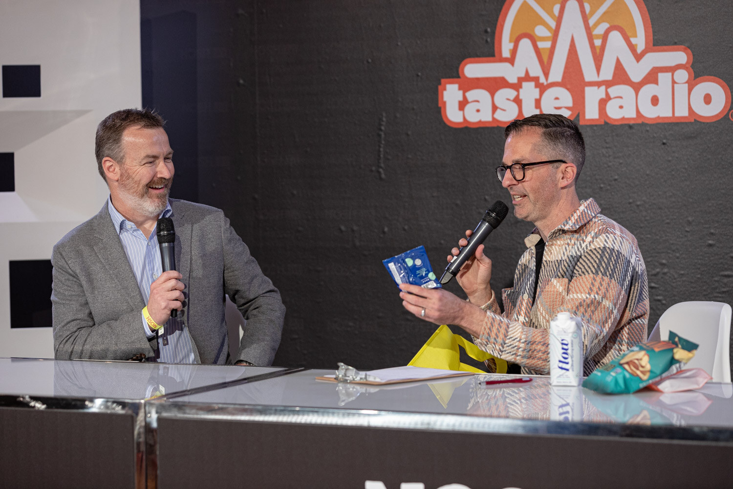 Man in a gray blazer laughing beside a man in glasses and a plaid jacket holding a blue package at the studio desk.