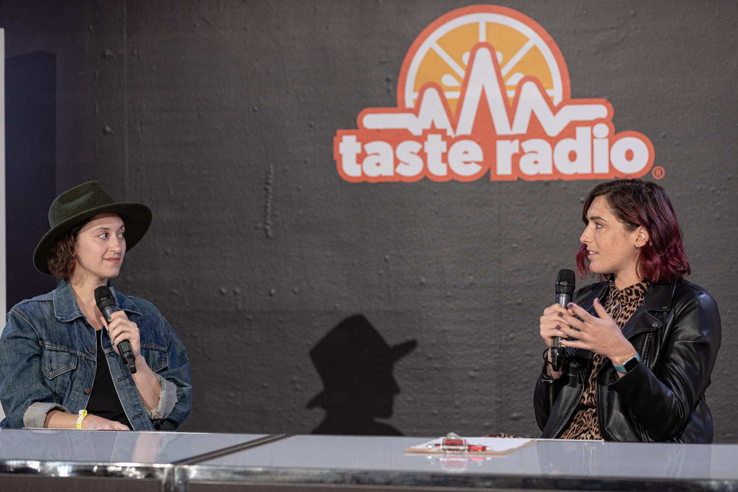 Woman in a green hat and denim jacket and woman in a black leather jacket with mics at the Taste Radio desk.