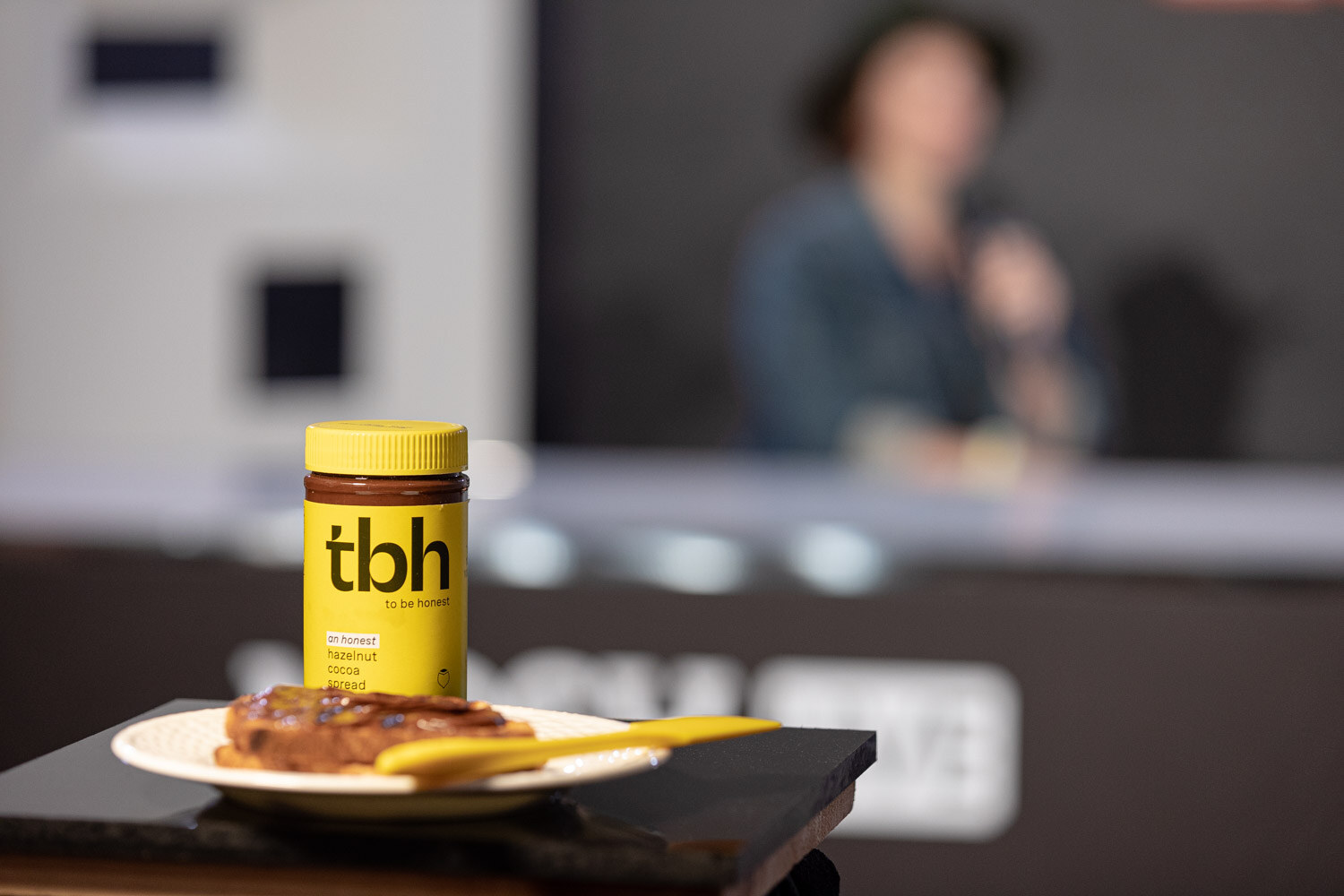 Yellow jar of tbh spread beside a plate of toast with a blurred interview behind it.