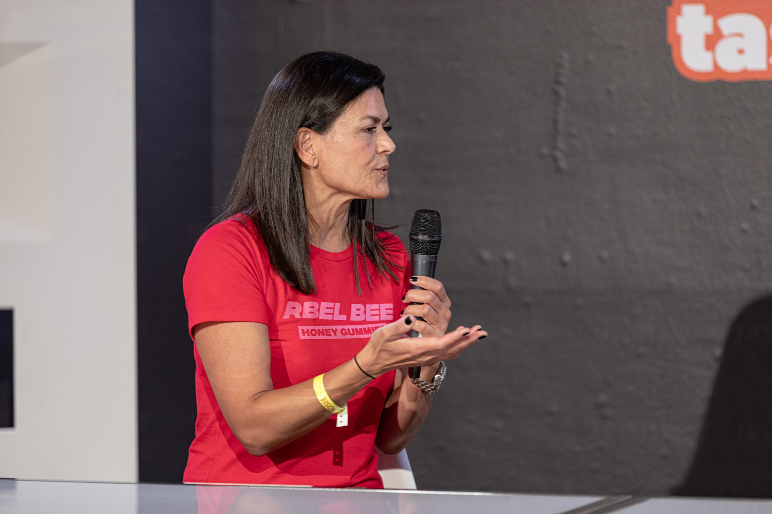 Woman in a red Rebel Bee Honey Gummies t-shirt holding a mic, one hand open.