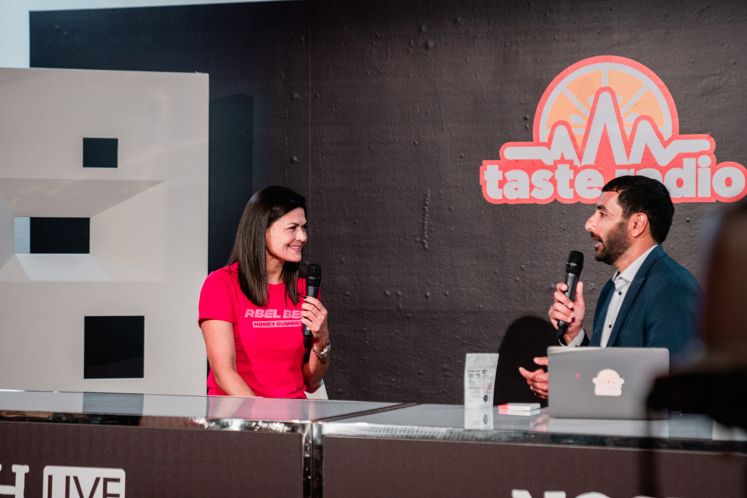 Woman in a pink Rebel Bee shirt and man in a navy blazer with mics at the Nosh Live desk.