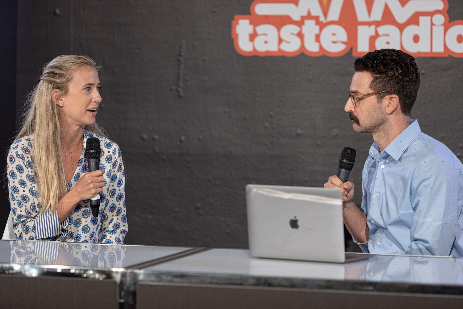 Blond woman in a blue patterned blouse and a man in a blue shirt with mics beside a laptop.